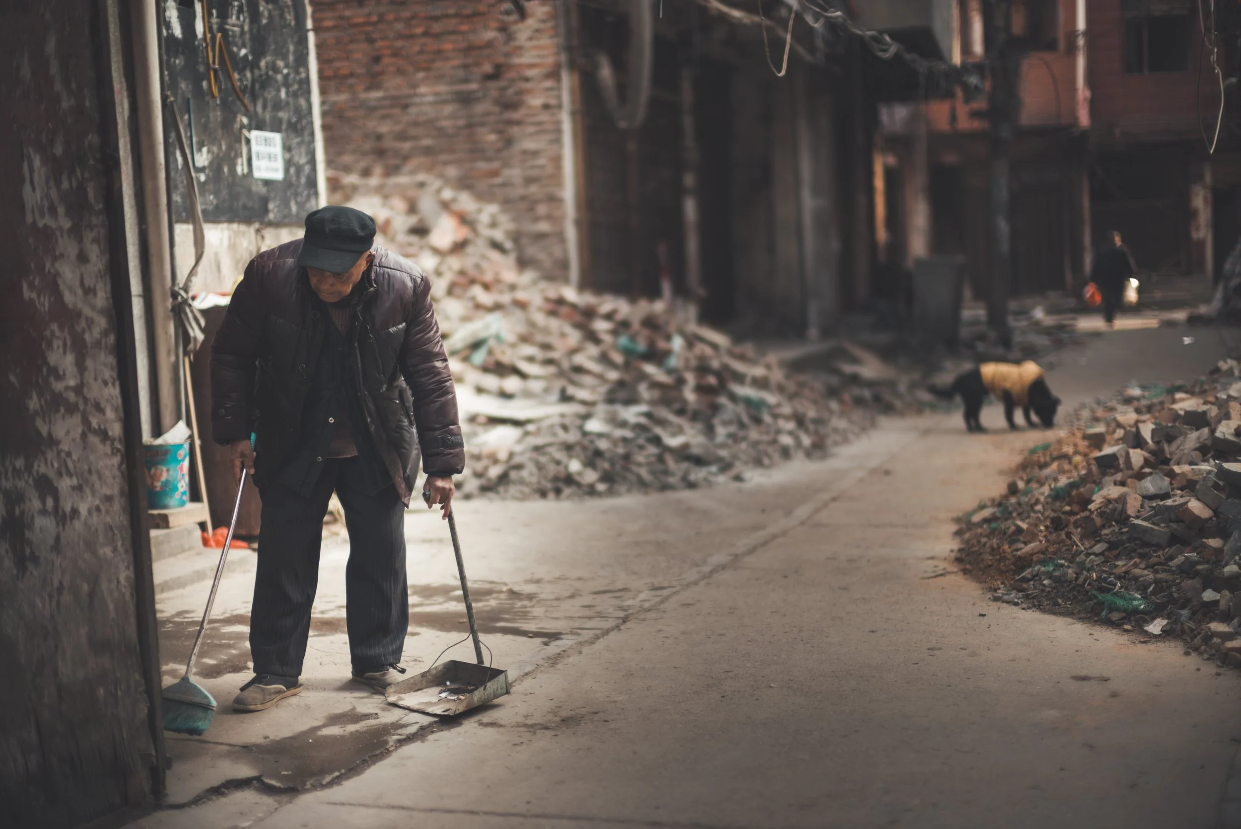 An elderly man with a hat and jacket sweeping the sidewalk with a broom and dustpan, with construction debris and partially built brick walls in the background.