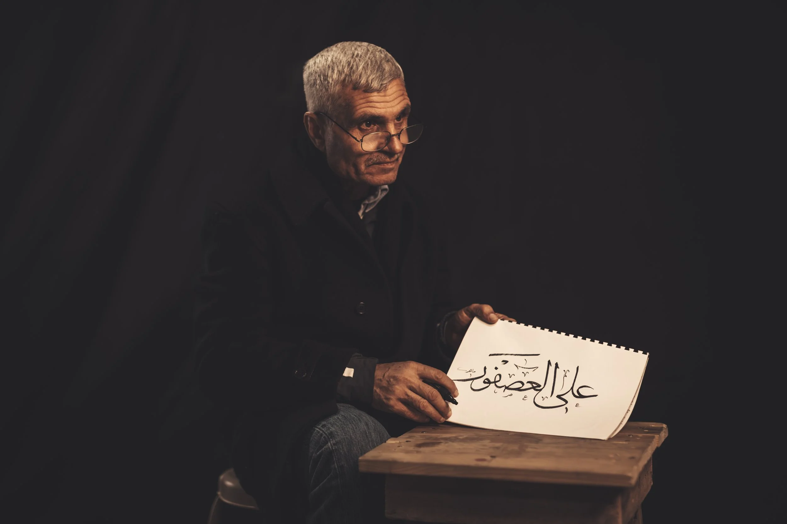 An older man with glasses and gray hair is sitting at a wooden table, holding a large white sign with Arabic writing on it. The background is dark.