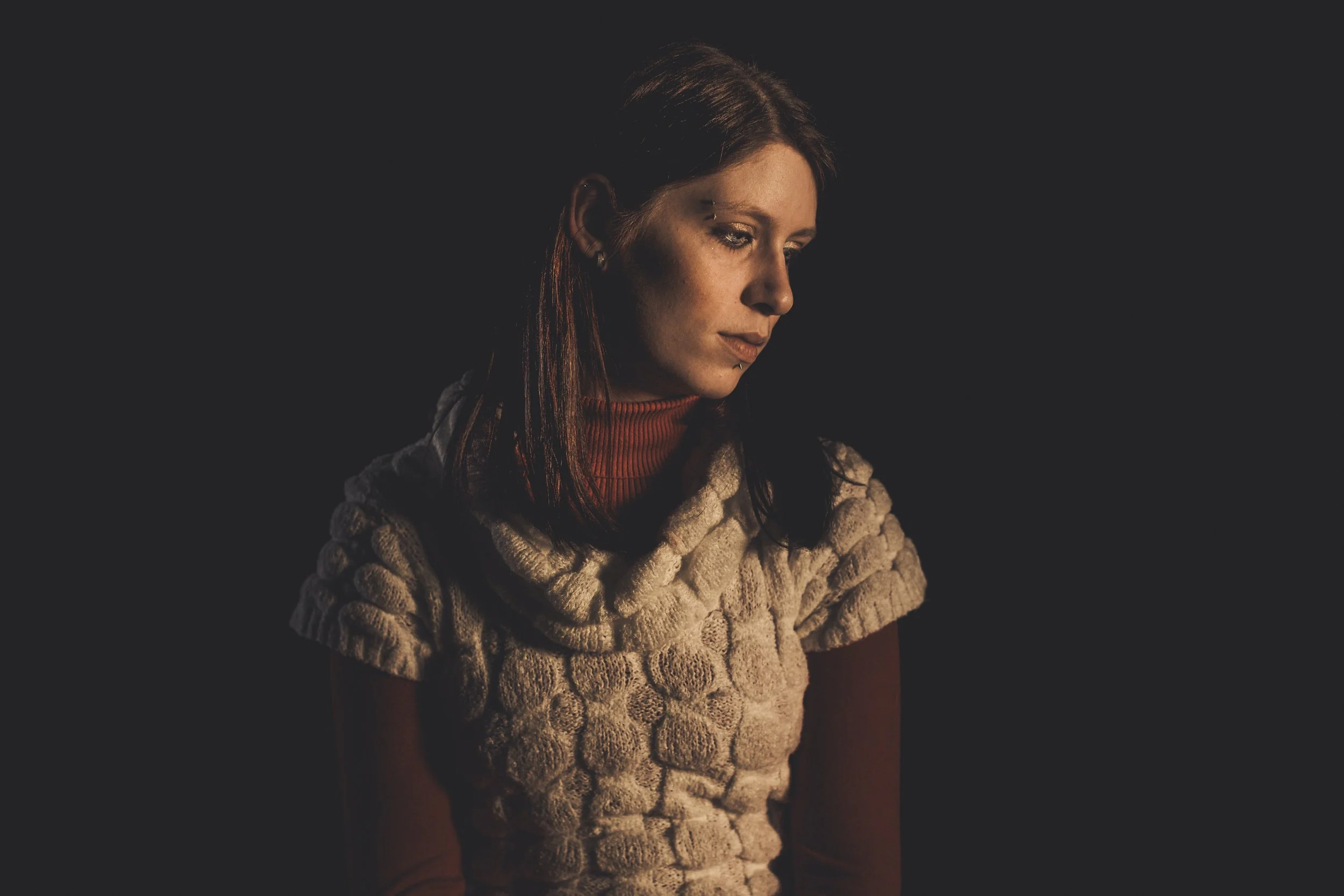 A woman with dark hair and light skin wearing a cozy, textured beige sweater with a high collar, looking thoughtfully downward against a dark background.