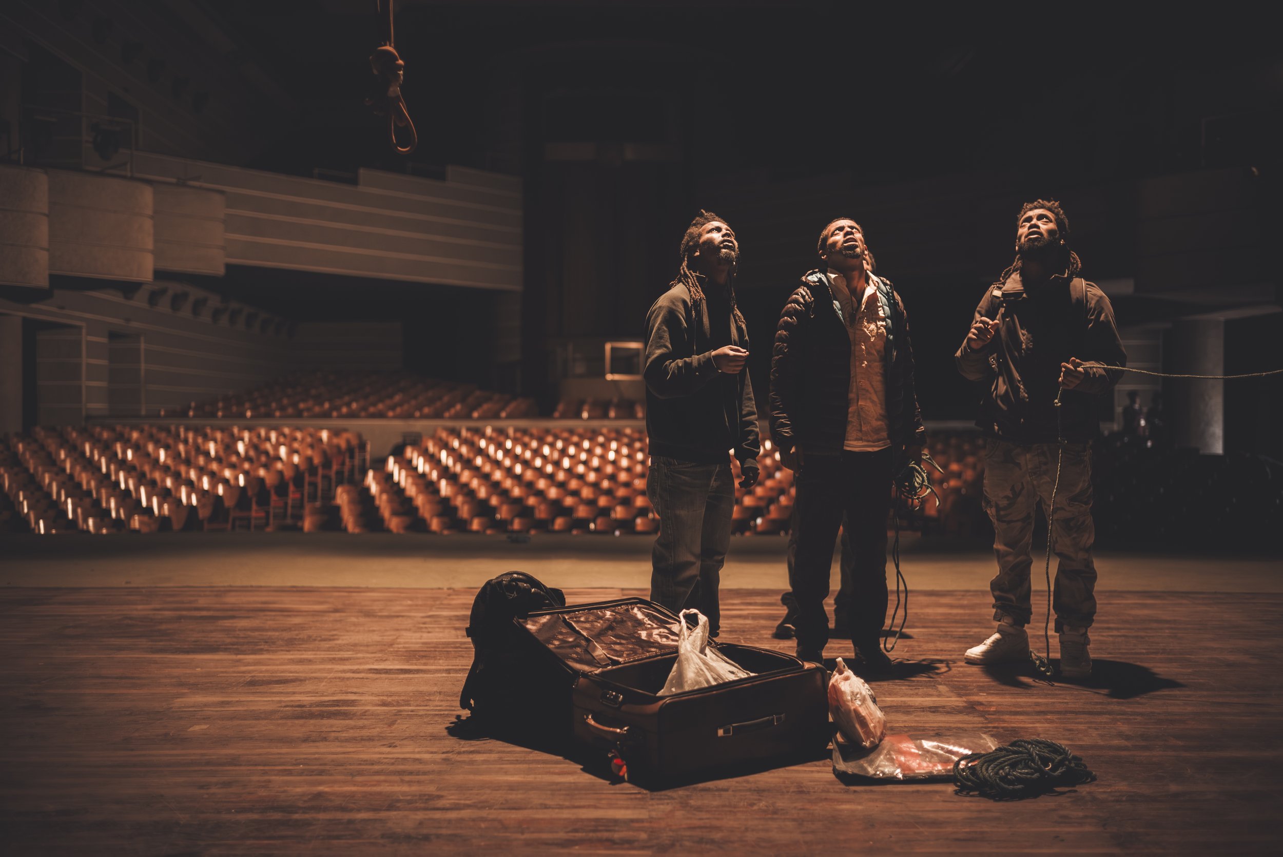 Three men with backpacks and work gloves looking up inside an empty theater or auditorium with wooden stage, orange seats, and dim lighting, surrounded by open luggage containing clothes, ropes, and bags.