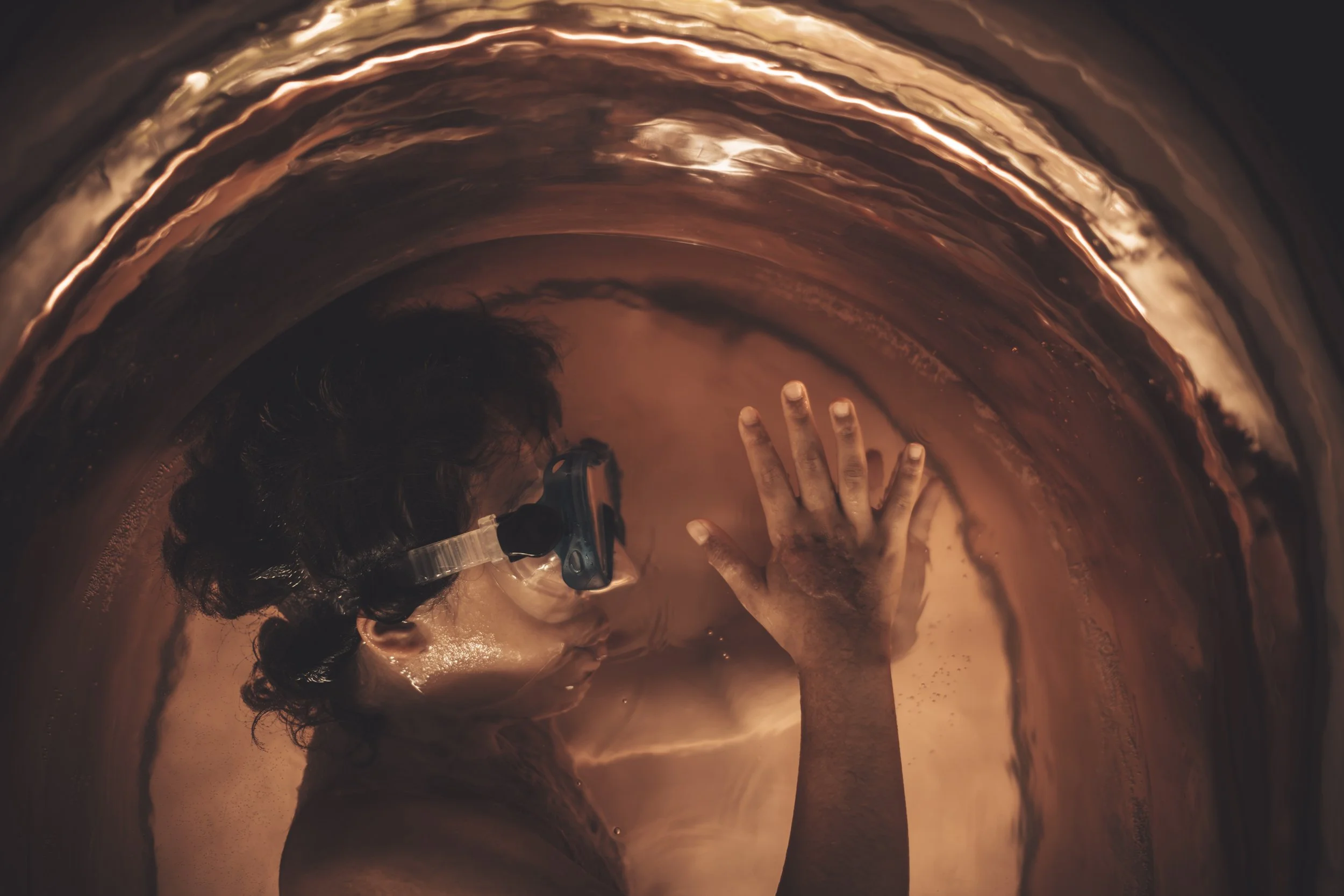 Person submerged in a copper-toned tub with their hand resting on the side, wearing goggles and surrounded by reflective, shimmering water.
