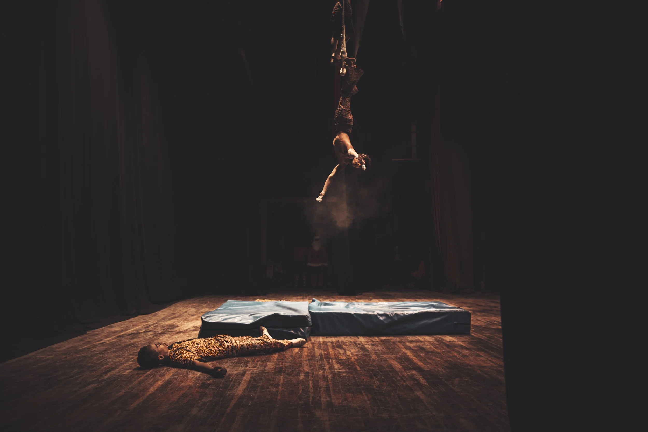 A person lying on a wooden floor with arms extended, while another person is suspended upside down, hanging from a harness or ropes, in a dark setting with spotlights.