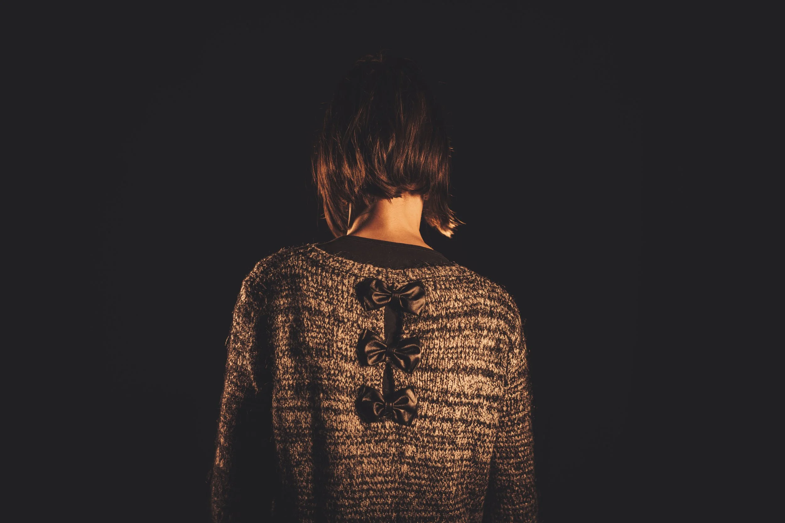 Rear view of a person with short, dark hair wearing a textured sweater with three black bows down the back, standing against a dark background.