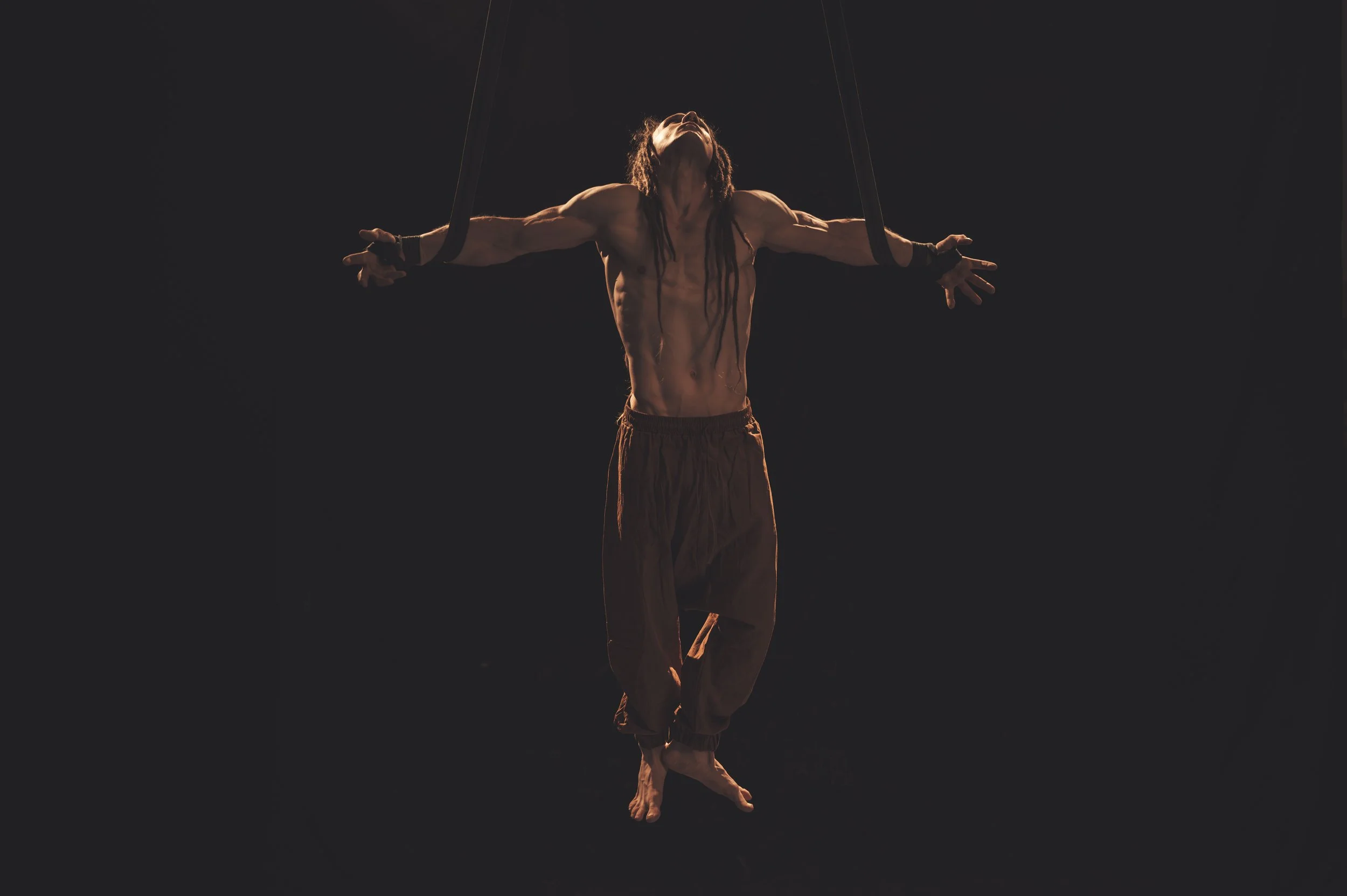 A muscular man with long dreadlocks performing aerial acrobatics using black straps, hanging against a dark background, illuminated with warm lighting.