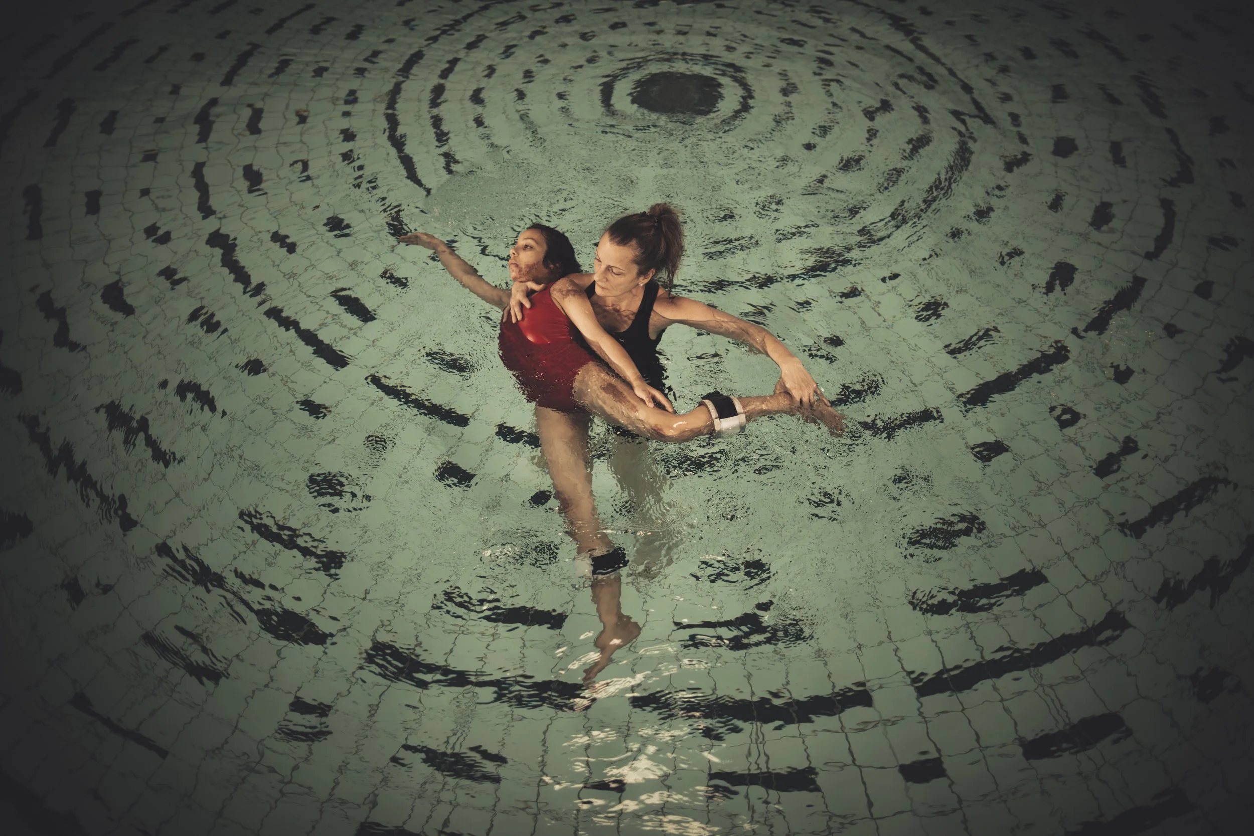 Two women swimming in a pool, embracing each other underwater, with ripples spreading outward from where they are holding each other.