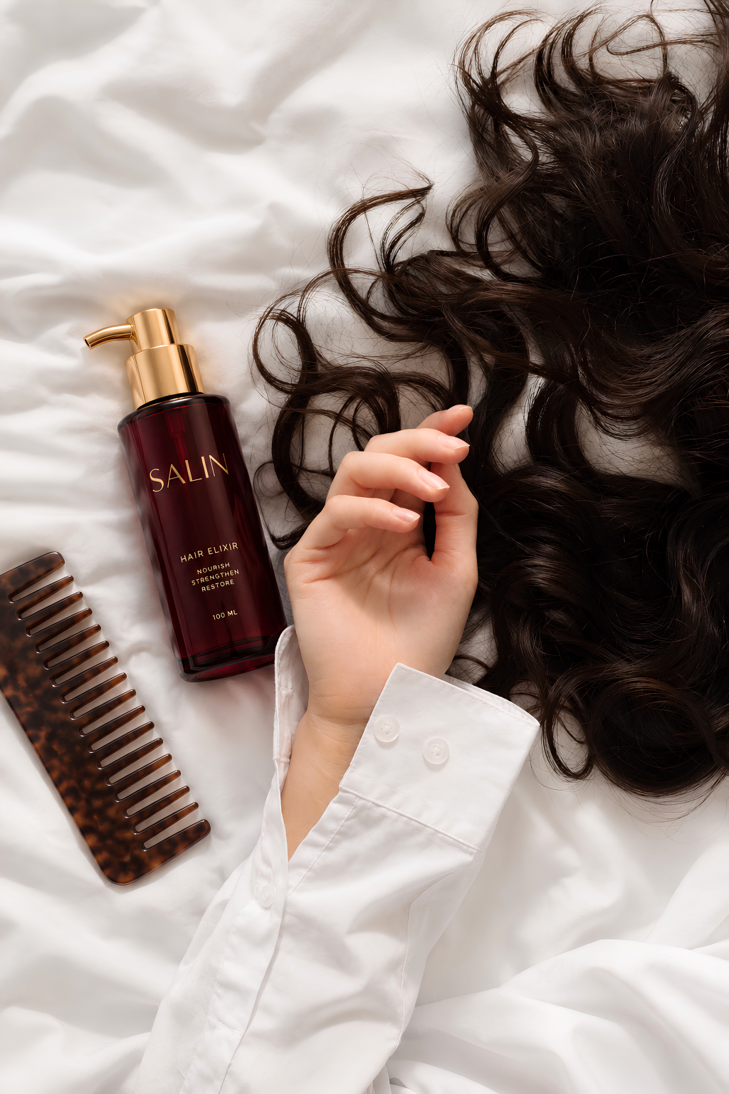 SALIN hair elixir bottle, wide-tooth comb, and hand resting on long dark hair against white bedding