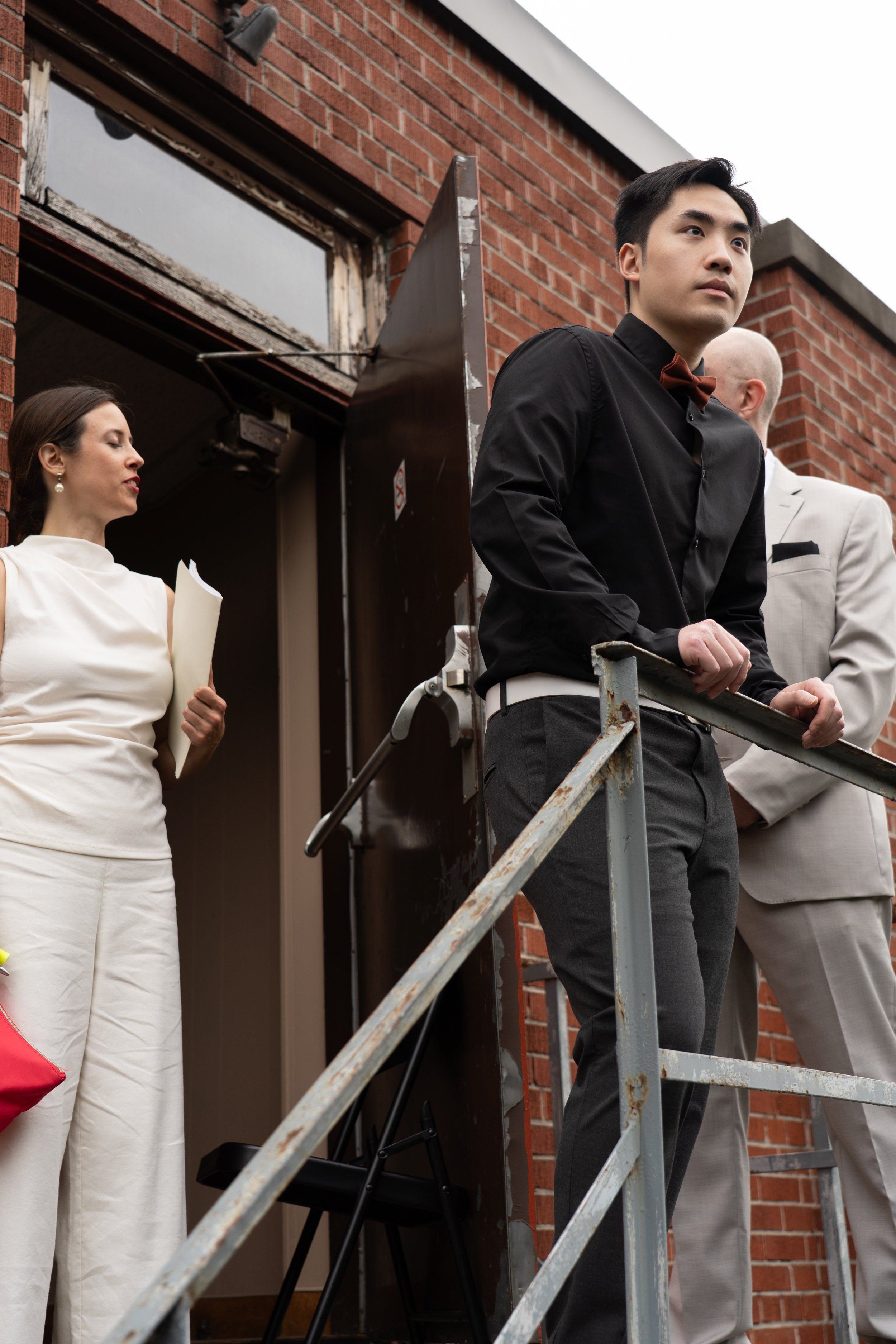 Three formally dressed people stand outside a brick building beside metal scaffolding, with a seated man in a black shirt and bow tie in the foreground.