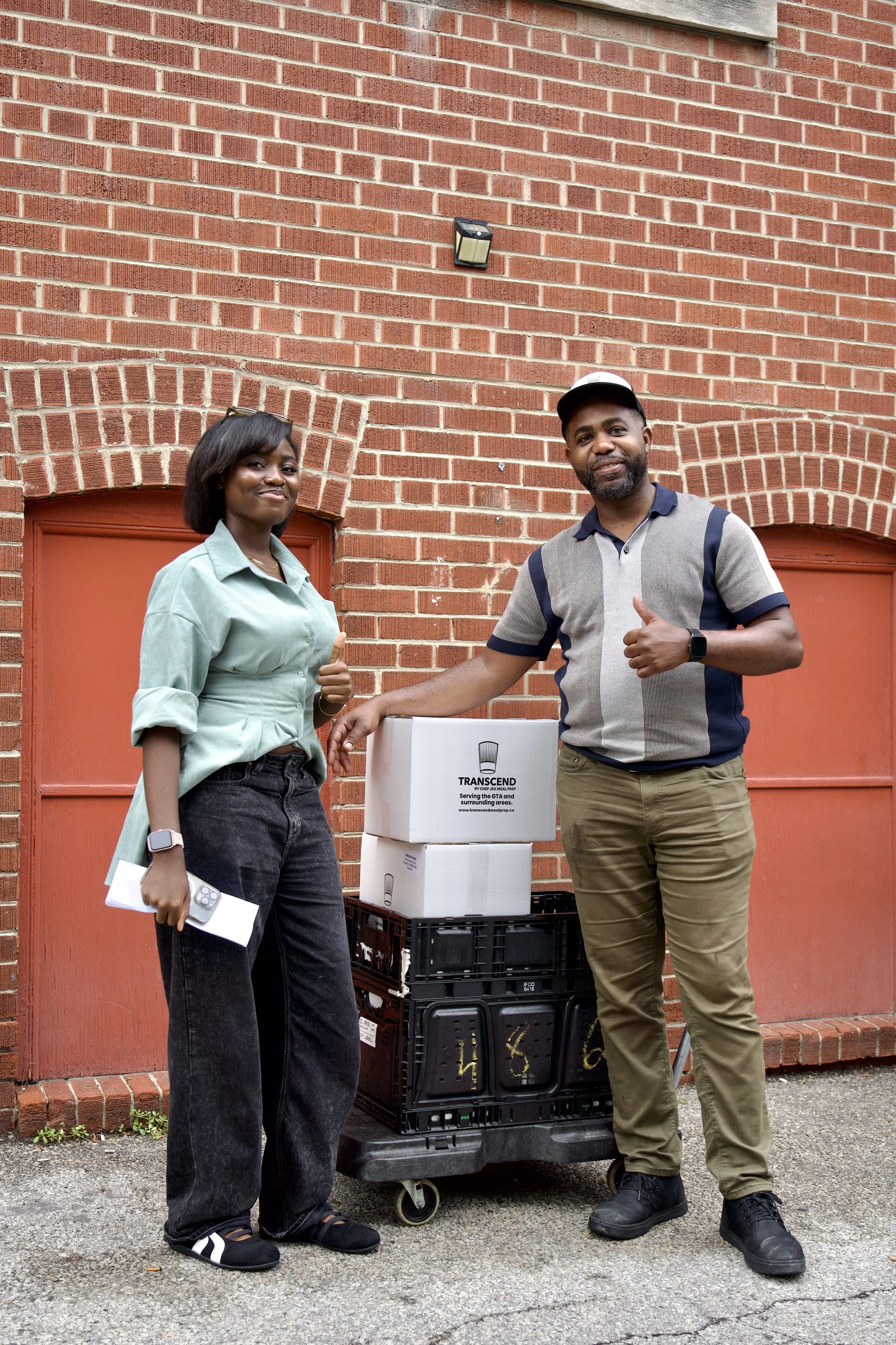 Two people giving thumbs-up beside stacked Transcend meal-preparation delivery boxes outside a brick building.