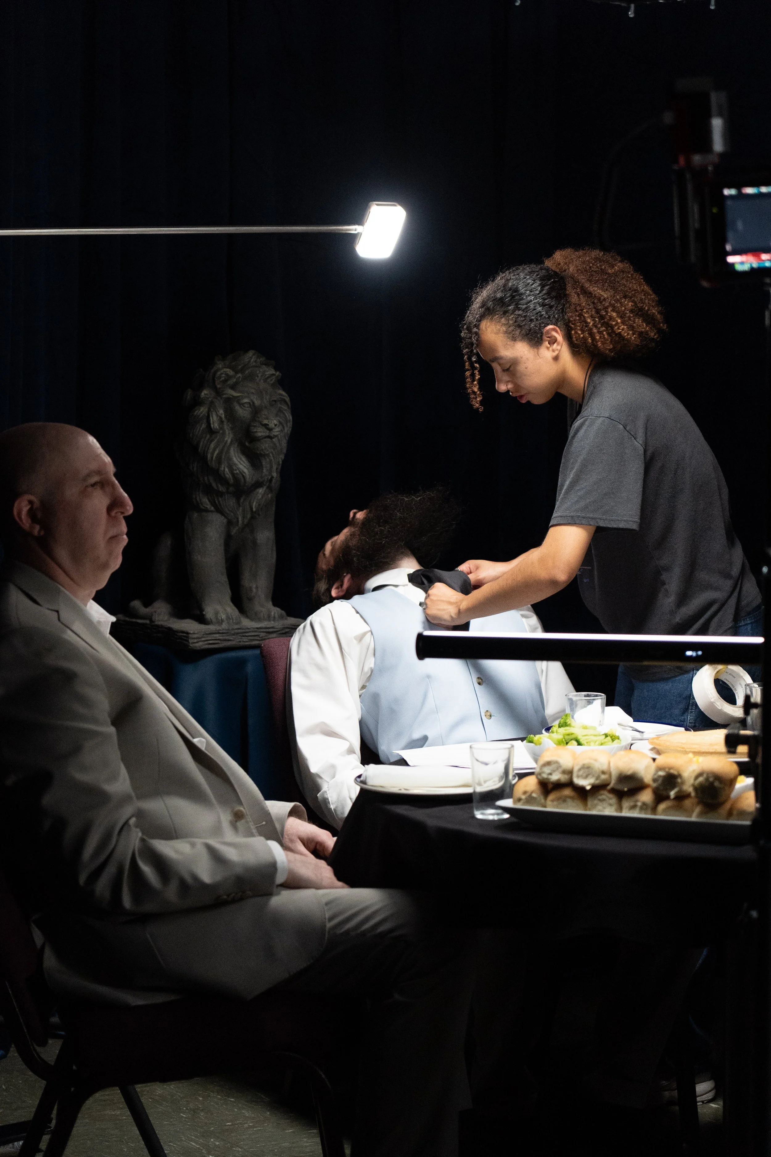 Theater backstage scene with an actor in costume, a performer assisting him, studio lights, a lion statue, and a meal on a table.