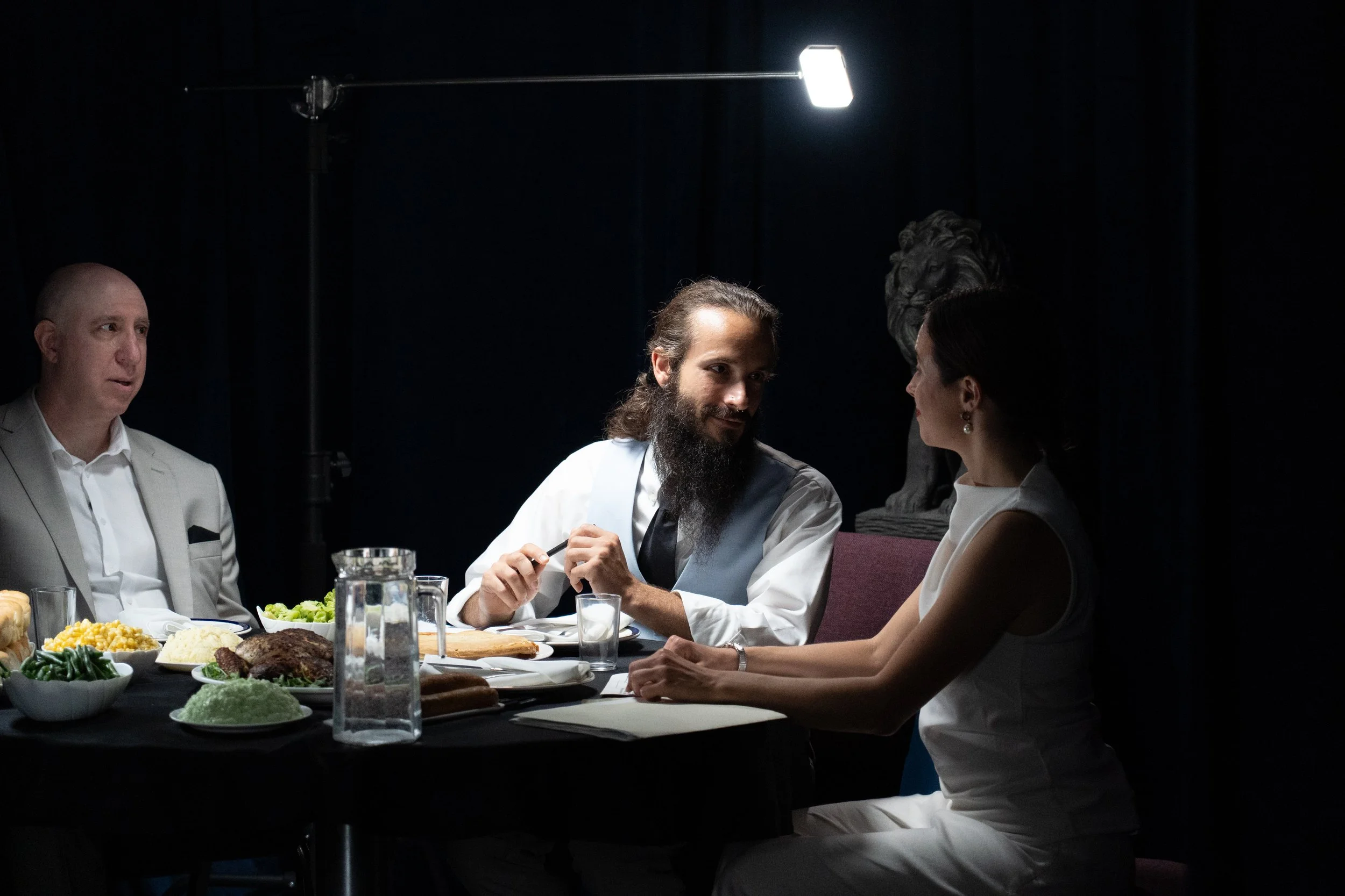 Three people dining at a table under dramatic lighting, with plates of food and a lion statue in the background