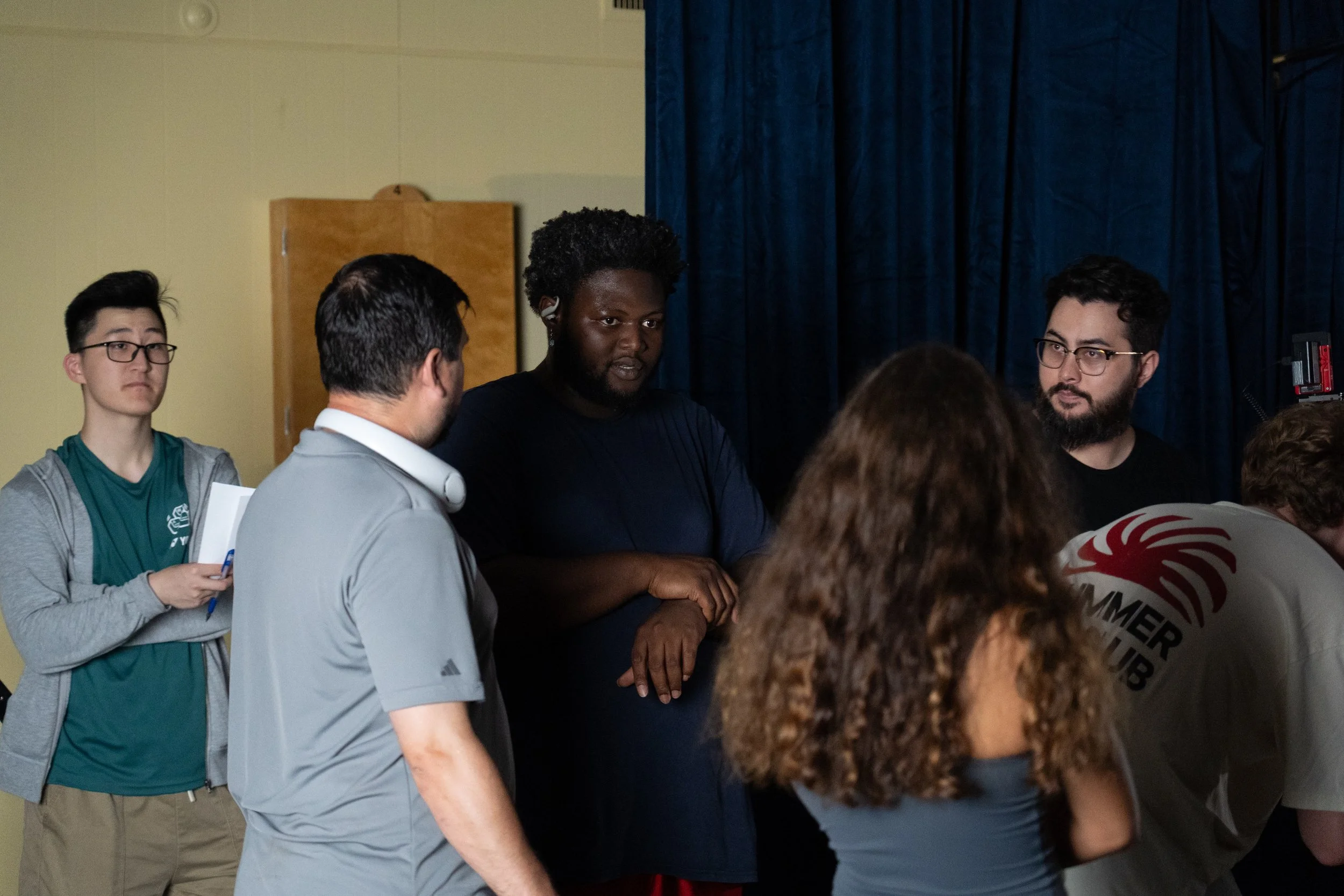 A diverse group of people gathers indoors near a curtained stage, appearing to participate in a discussion.