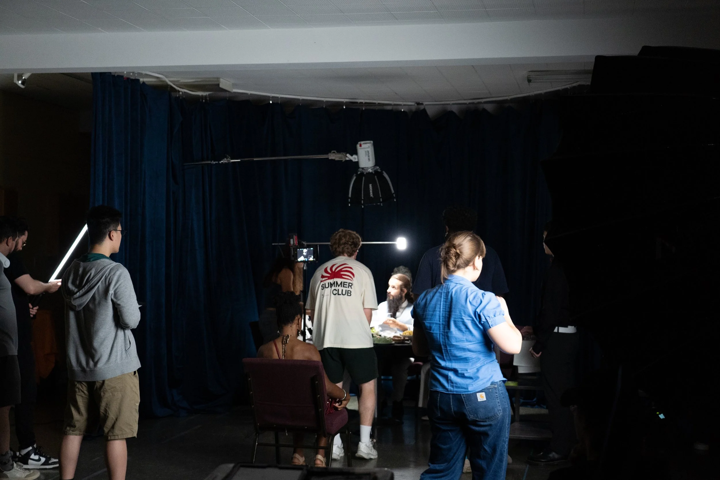 Film crew recording a dining scene on a dark set with studio lights, cameras, and blue curtains