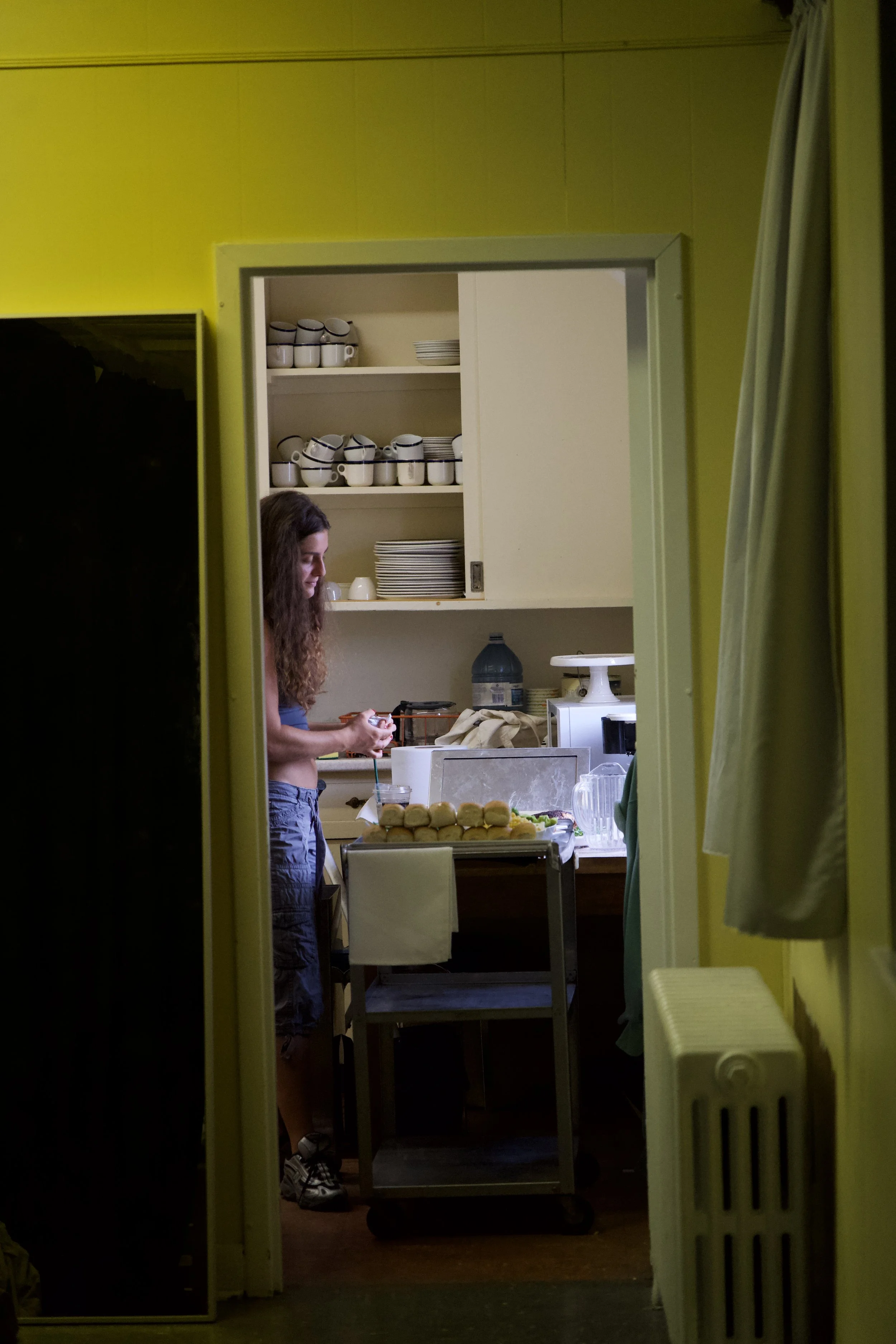 Person preparing food in a kitchen with shelves of cups and plates, viewed through an open doorway