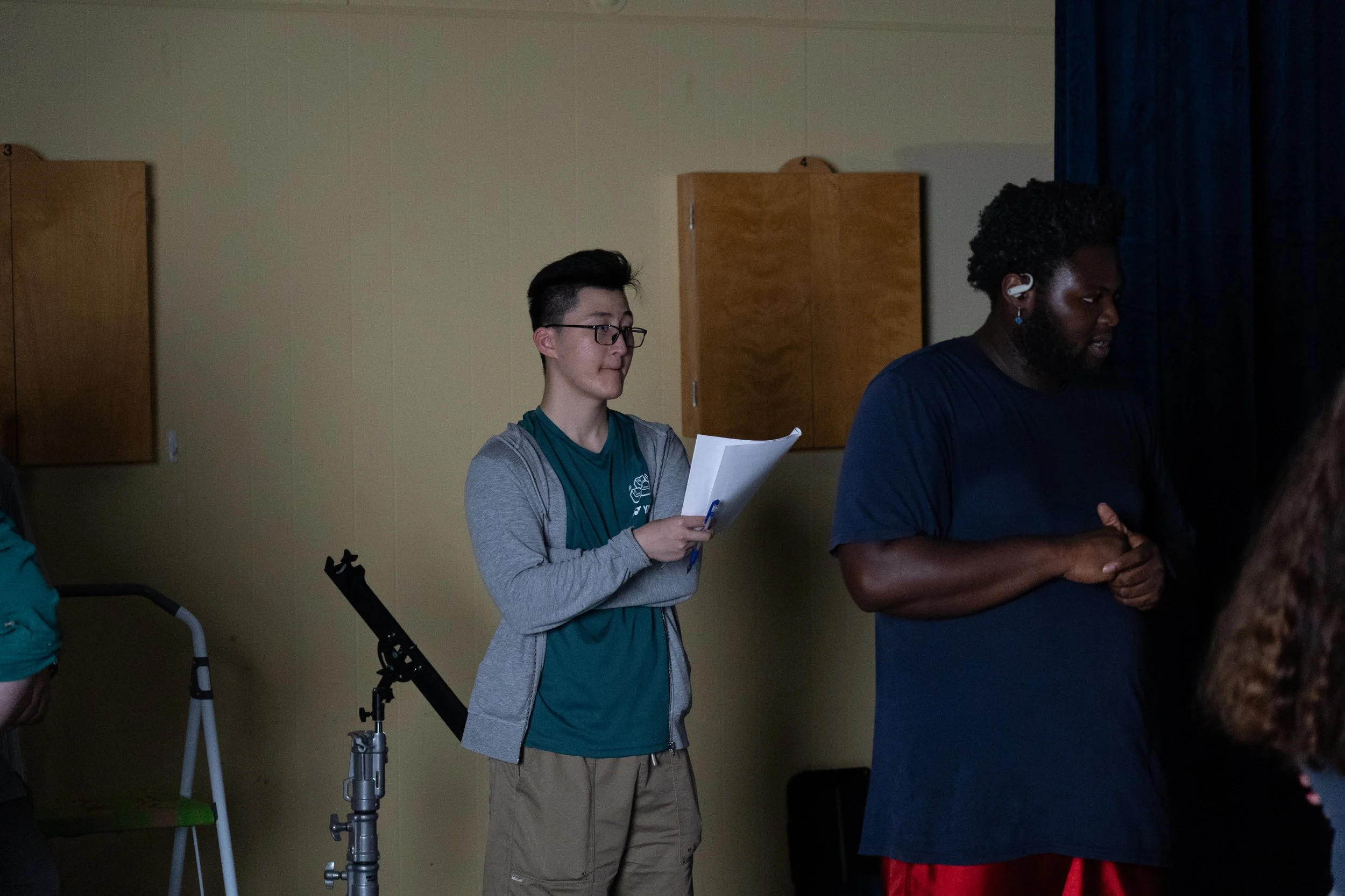 People rehearsing indoors, with one person holding a script beside a camera tripod.