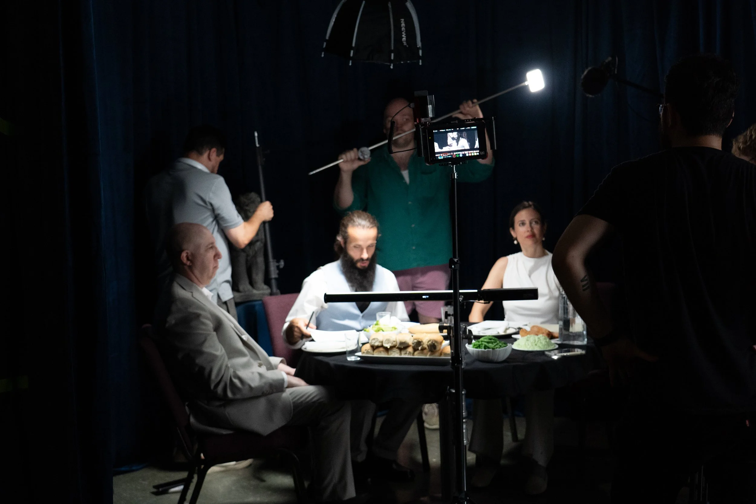 Film crew shooting a dinner scene with actors seated at a table surrounded by cameras and lighting equipment