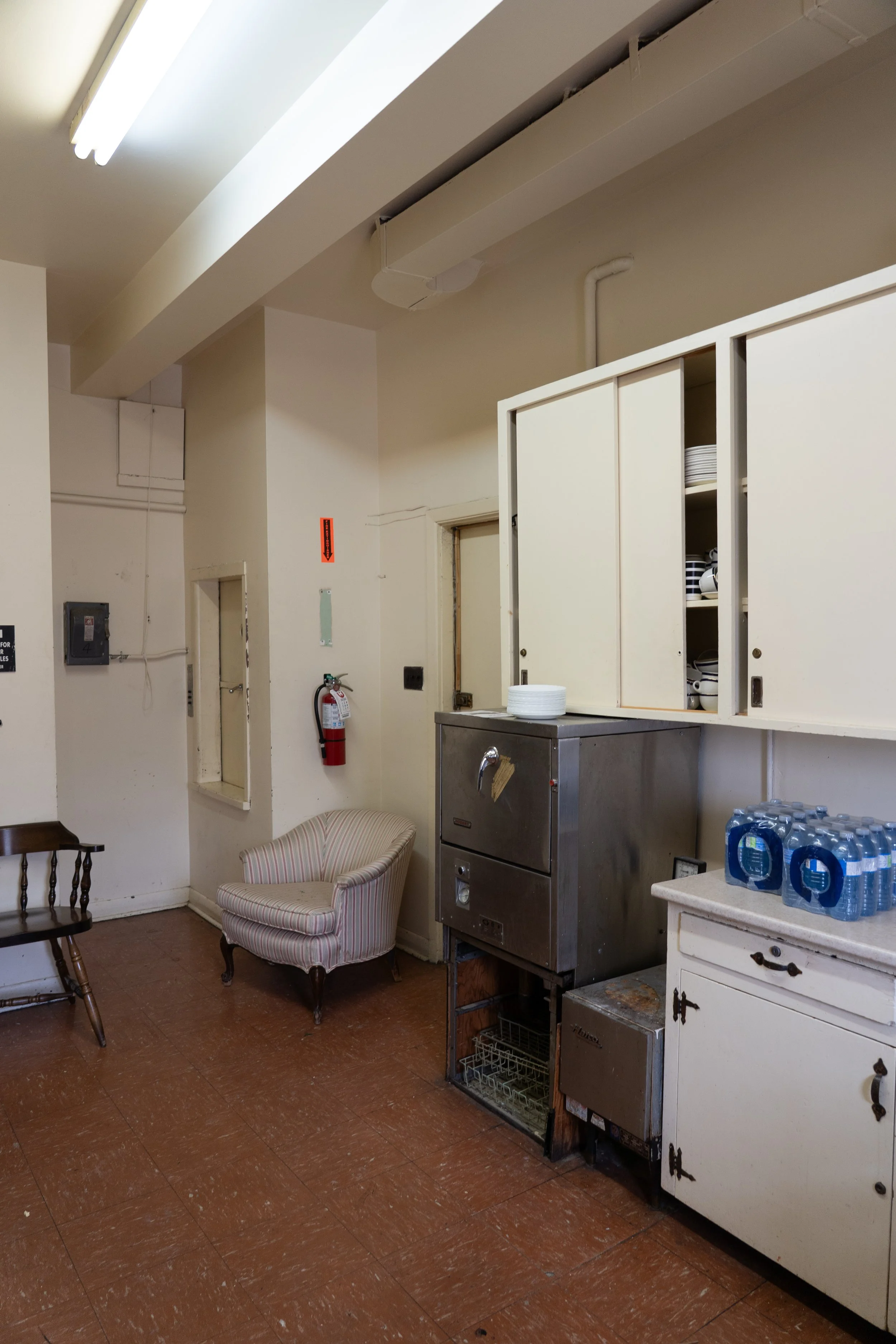 Institutional kitchen and dining area with open cabinets, stacked plates, commercial dishwasher, water bottles, fire extinguisher, and upholstered chair