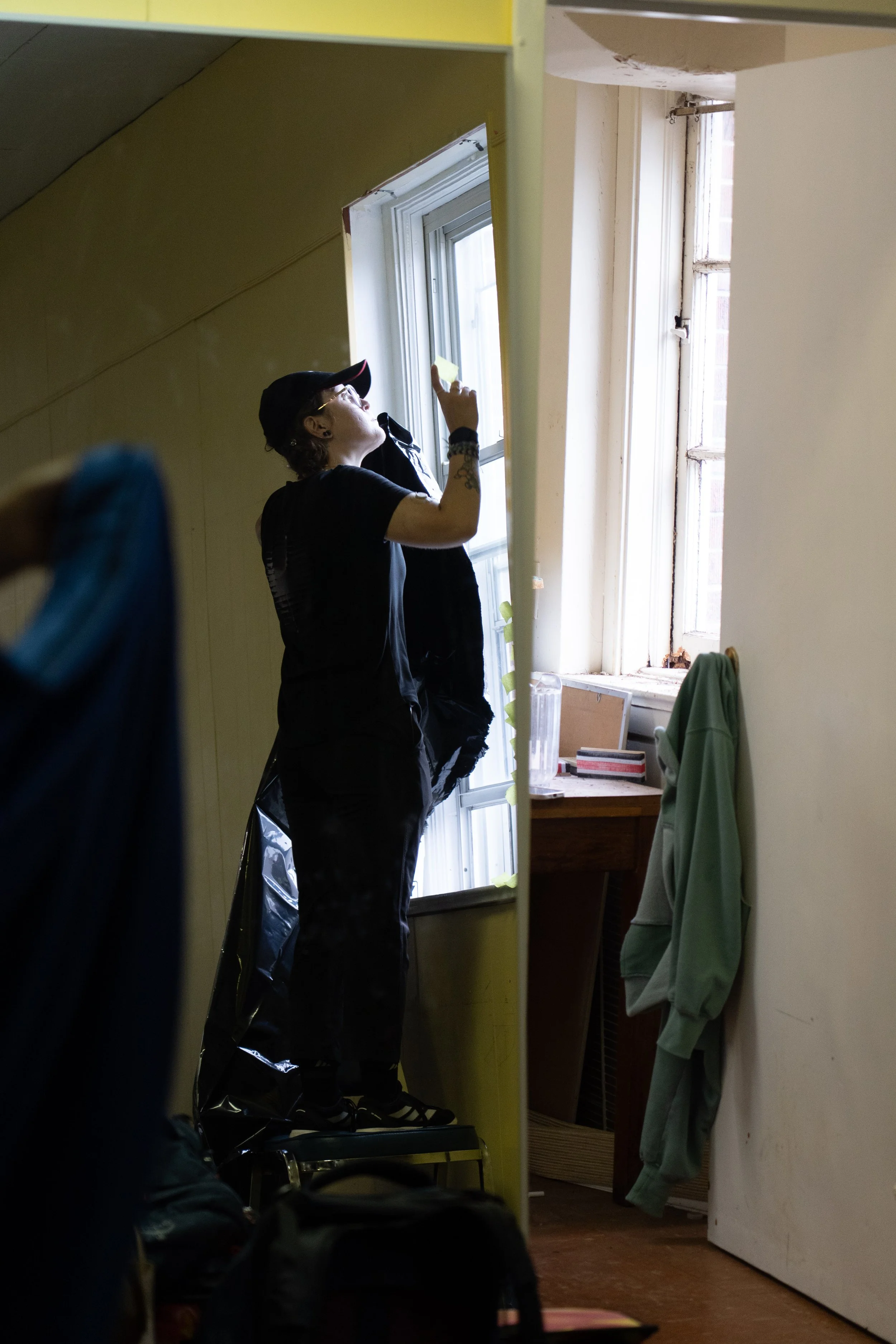 Person in a black costume and cap examines a window frame inside a room.