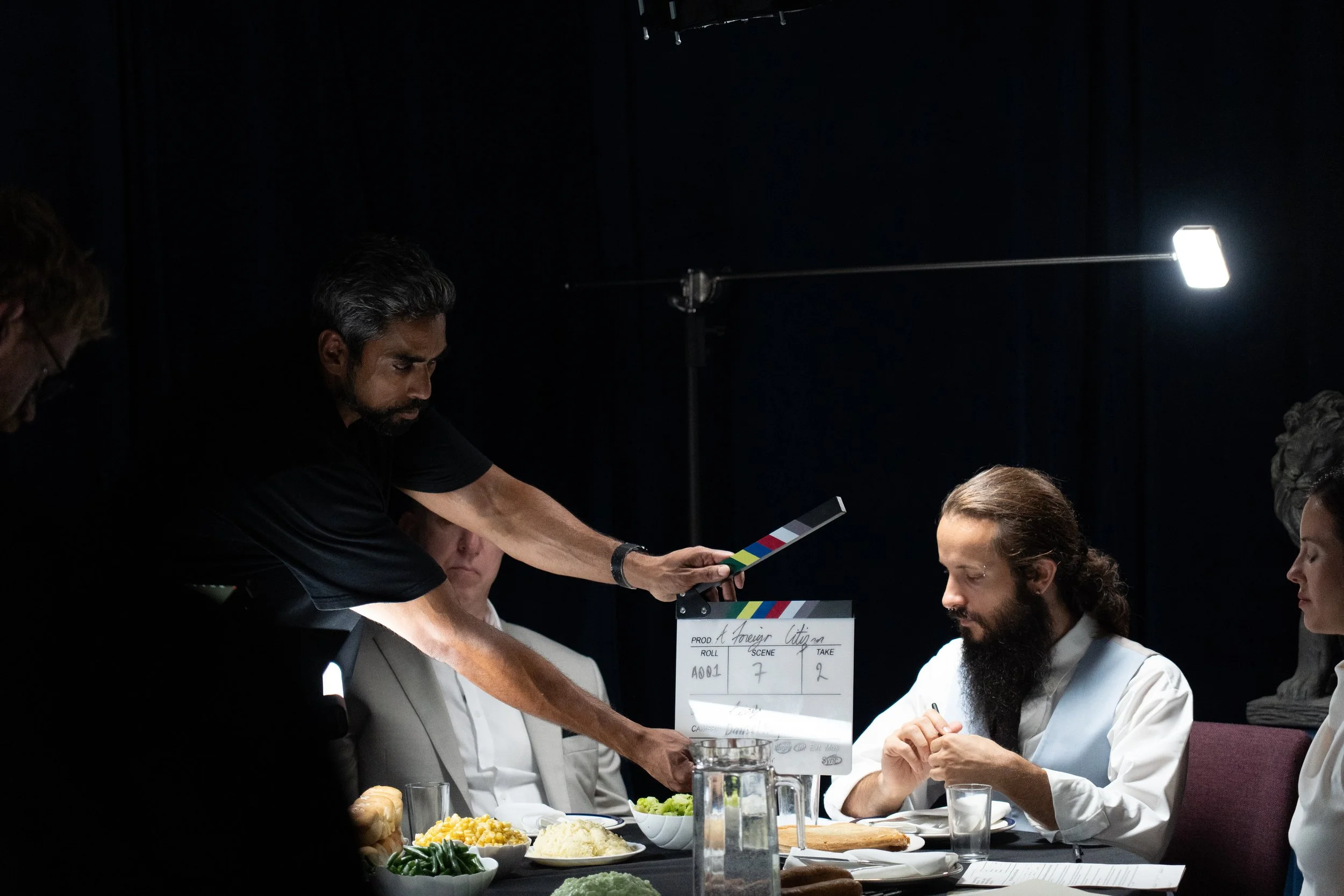 Film crew claps a slate while actors sit at a dinner table on a dark movie set.