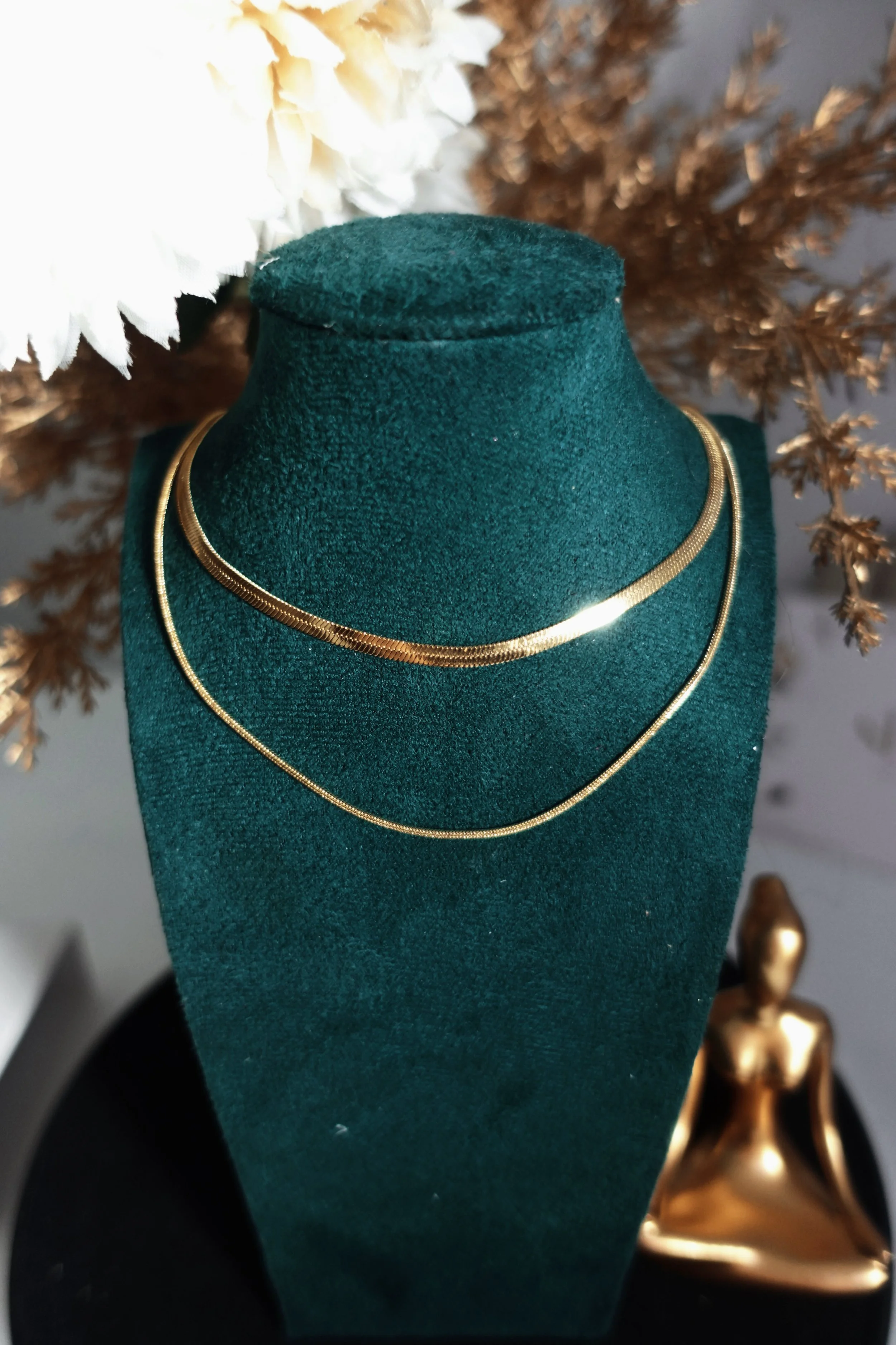Gold layered chain necklaces displayed on a teal velvet jewelry bust with white and bronze dried flowers.