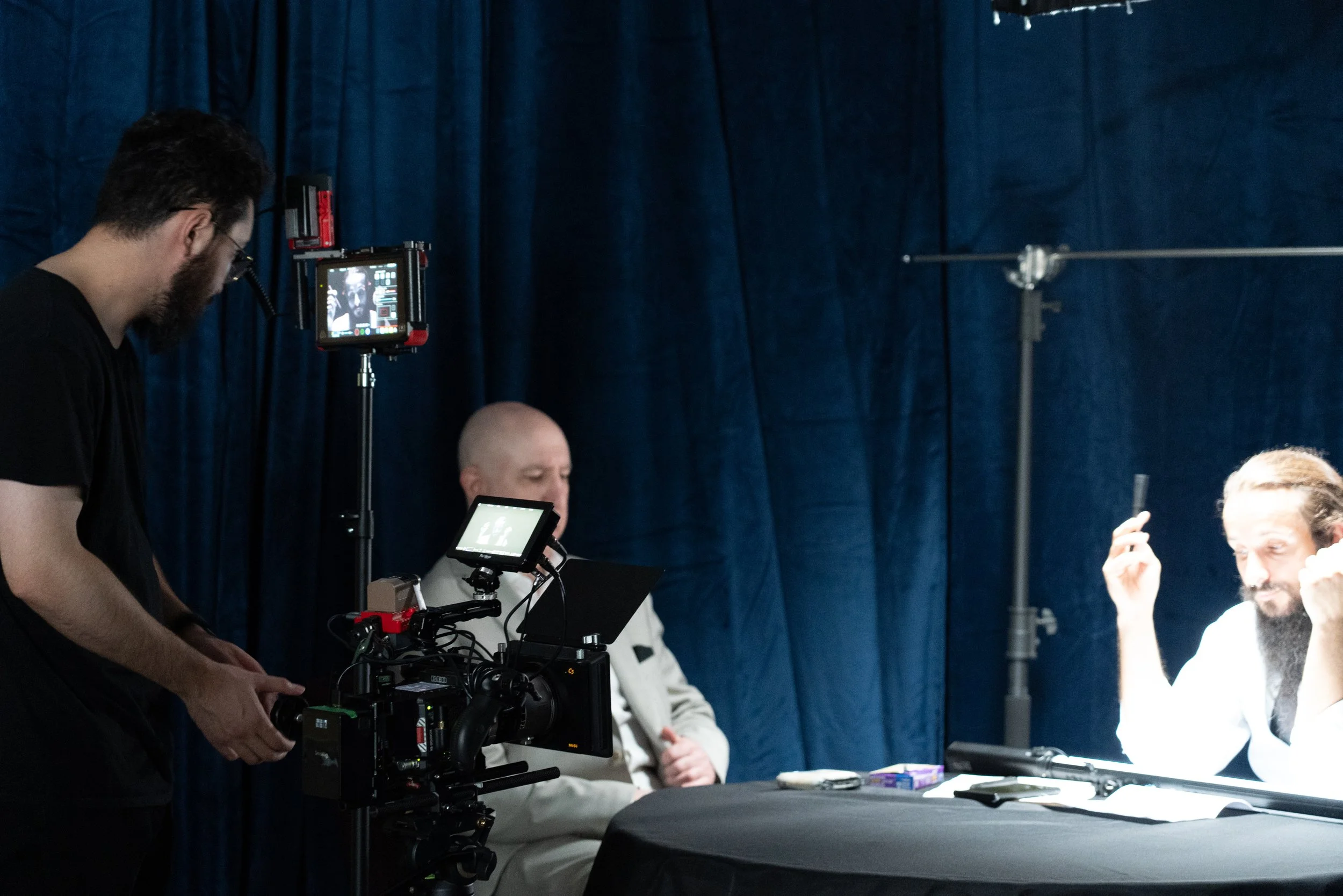 Film crew recording a bearded man seated at a table in front of blue curtains