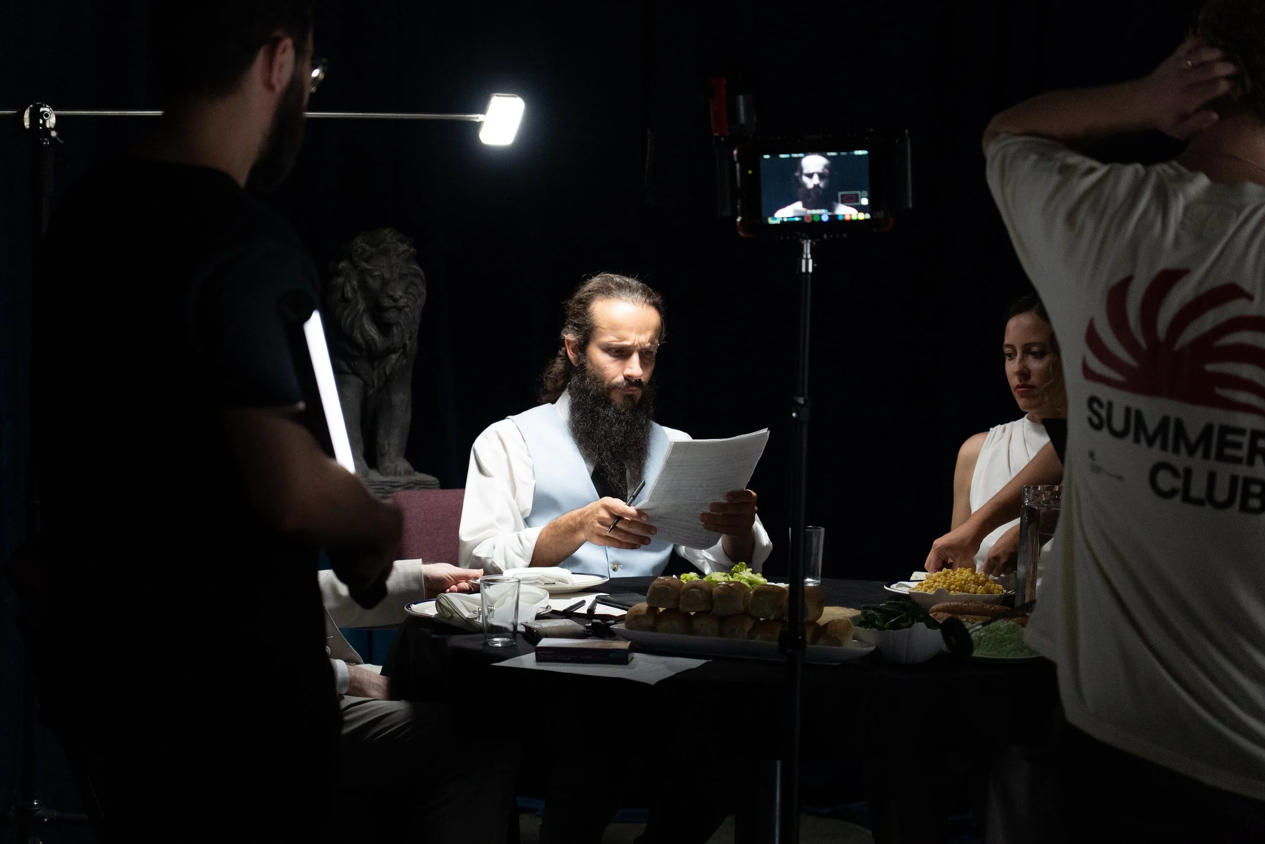 Behind-the-scenes film set with a bearded actor reading a script at a dinner table while crew members adjust lighting and camera equipment