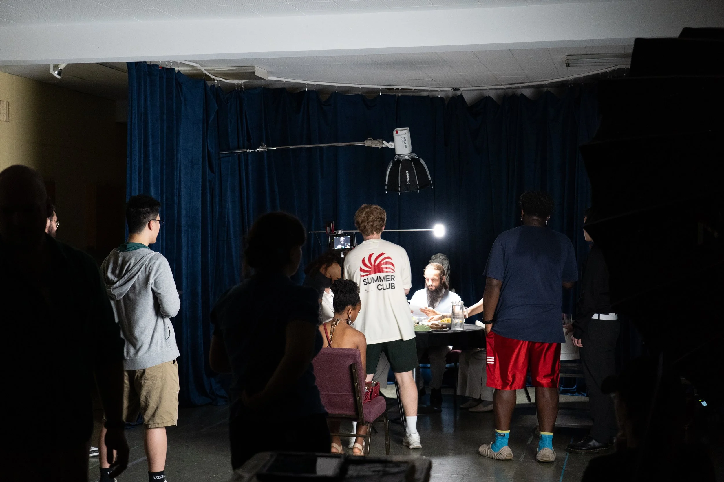 People gather around a dining table during a film shoot in a curtained room, with studio lights and a camera visible.