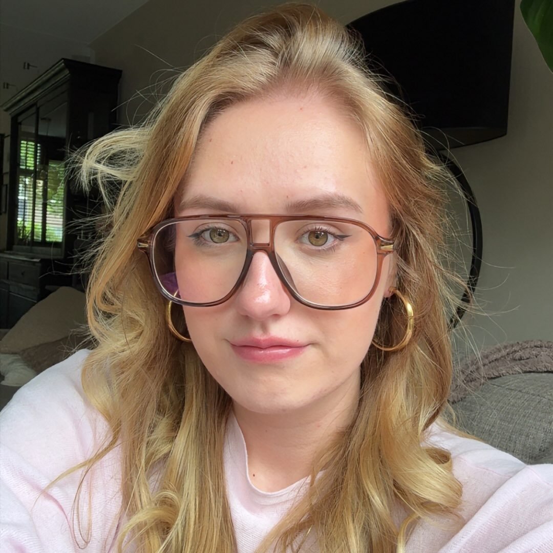 A young woman with blonde hair and light skin wearing large glasses and gold hoop earrings, sitting in a living room.