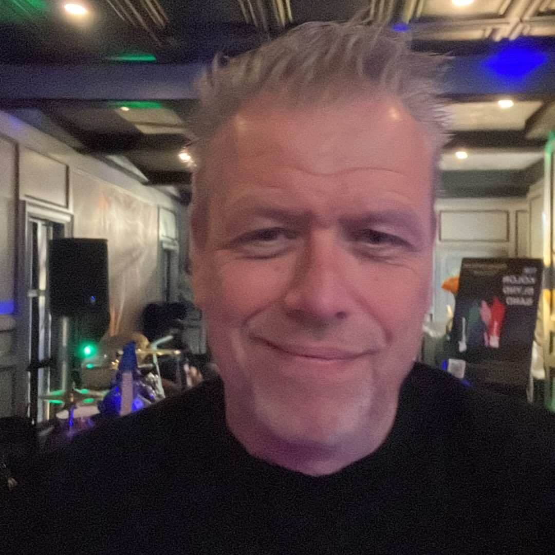 A middle-aged man with gray hair smiling in a dimly lit indoor setting, possibly at a bar or nightclub.