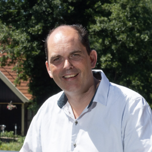 A smiling man with short dark hair, wearing a white shirt, standing outdoors with trees and a house in the background.