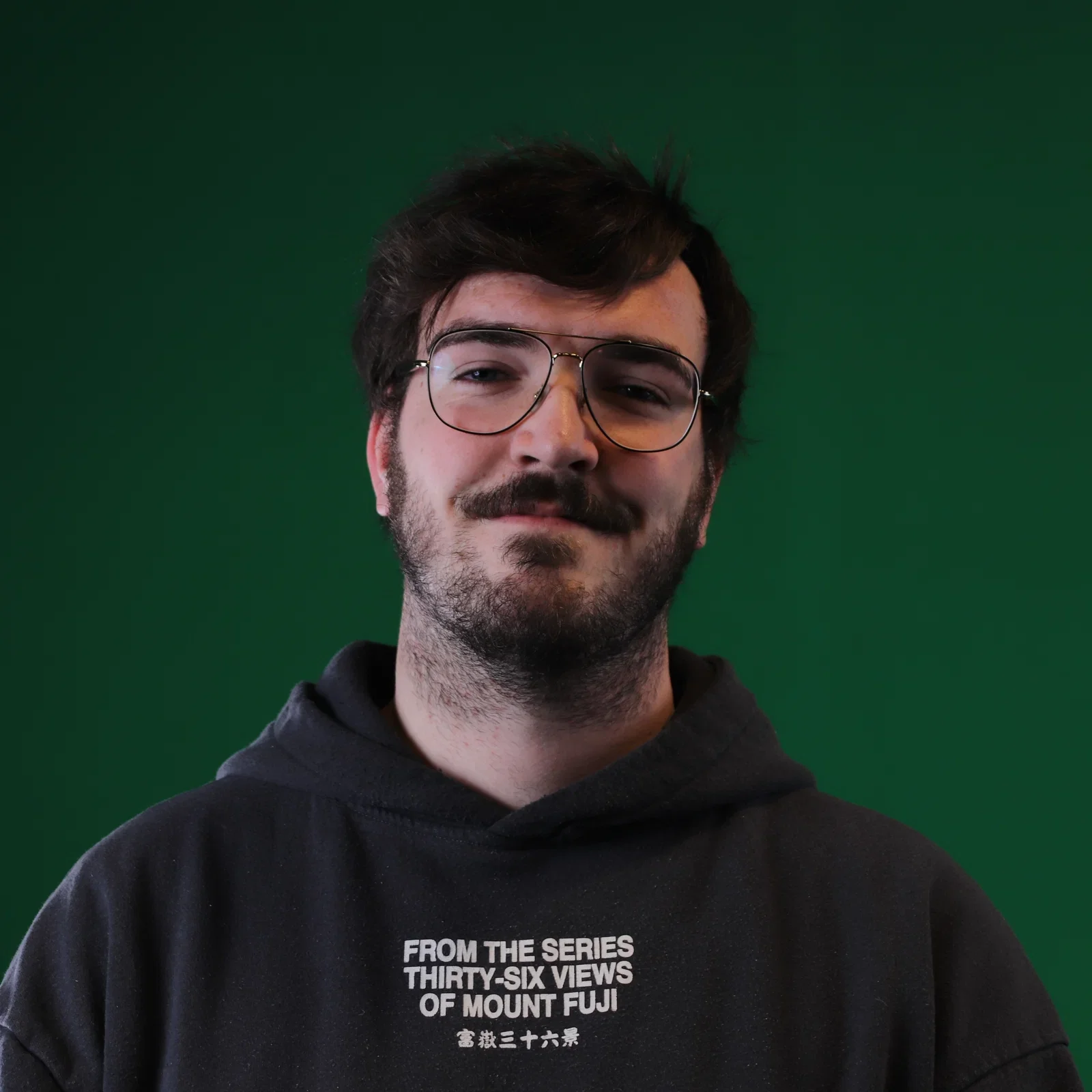 A man with dark brown hair, beard, and glasses wearing a black hoodie with white text on the front, standing in front of a solid green background.
