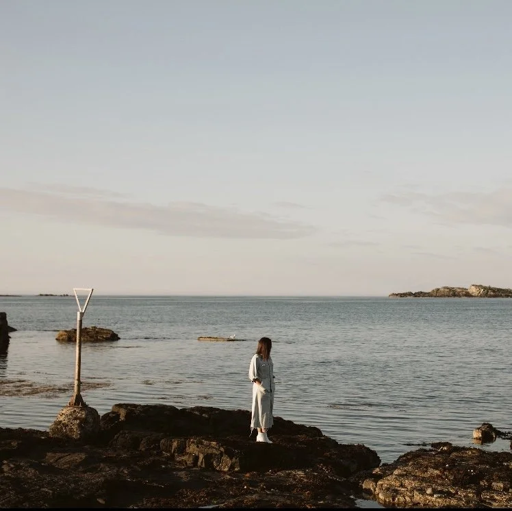 A woman stands on rocks by the ocean, looking at the water during sunset or sunrise, with a partly cloudy sky.