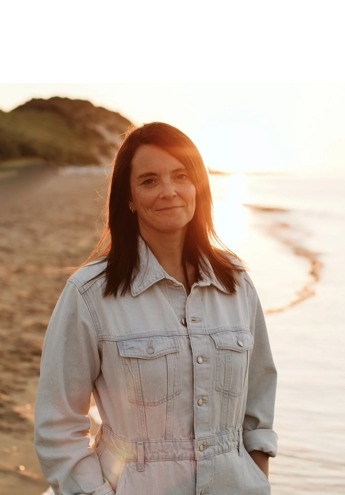 A woman with reddish hair standing on a beach at sunset, wearing a light denim jacket, with an ocean and green hills in the background.