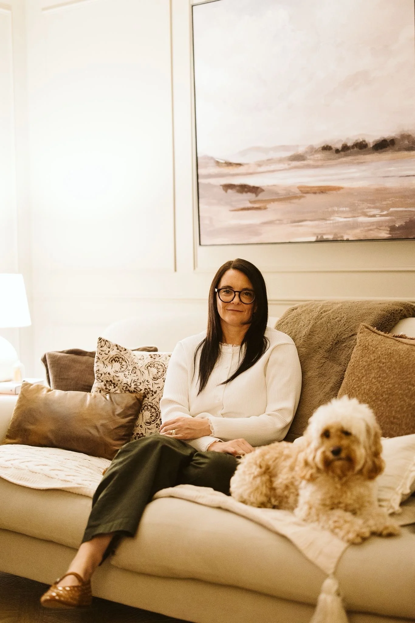 A woman with glasses sitting on a beige couch in a living room, with a fluffy dog lying in front of her, pillows behind her, and a large abstract landscape painting on the wall behind.