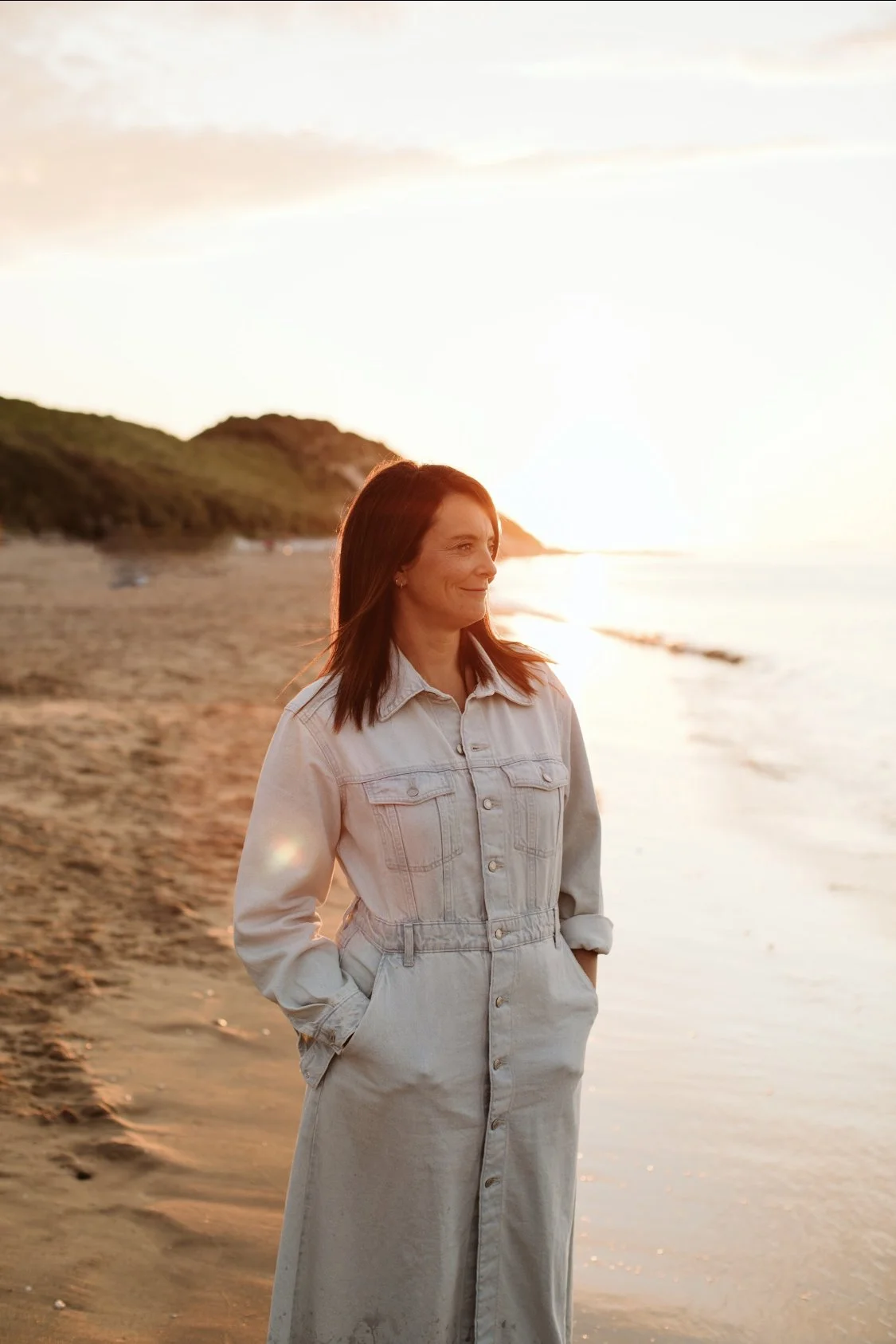 A woman with shoulder-length brown hair stands on the beach during sunset, wearing a light-colored denim jumpsuit with hands in pockets, overlooking the ocean with hills in the background.