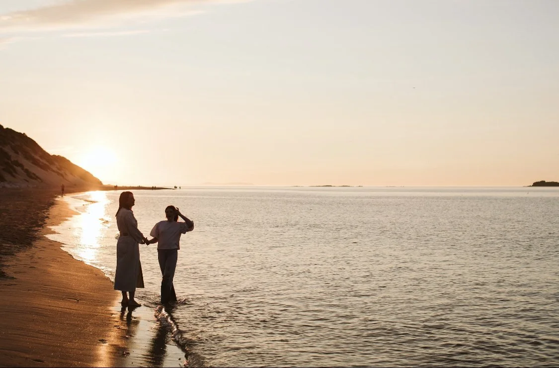 mother and daughter holding hands on a beach at sunset, with the ocean and a sandy hill in the background.