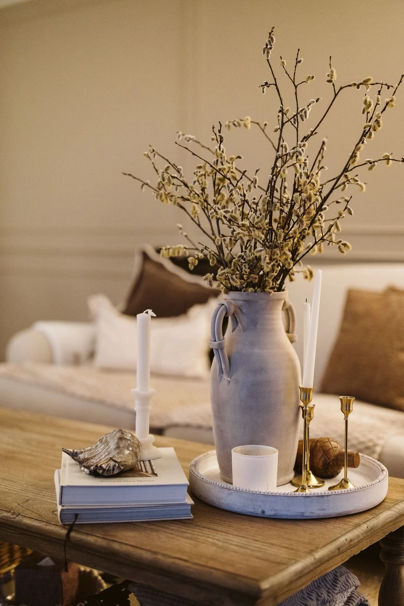 A white ceramic vase with dried pussy willow branches on a round tray, surrounded by white candles, seashells, and gold candlesticks on a wooden table, with a blurred background of a cozy indoor space.