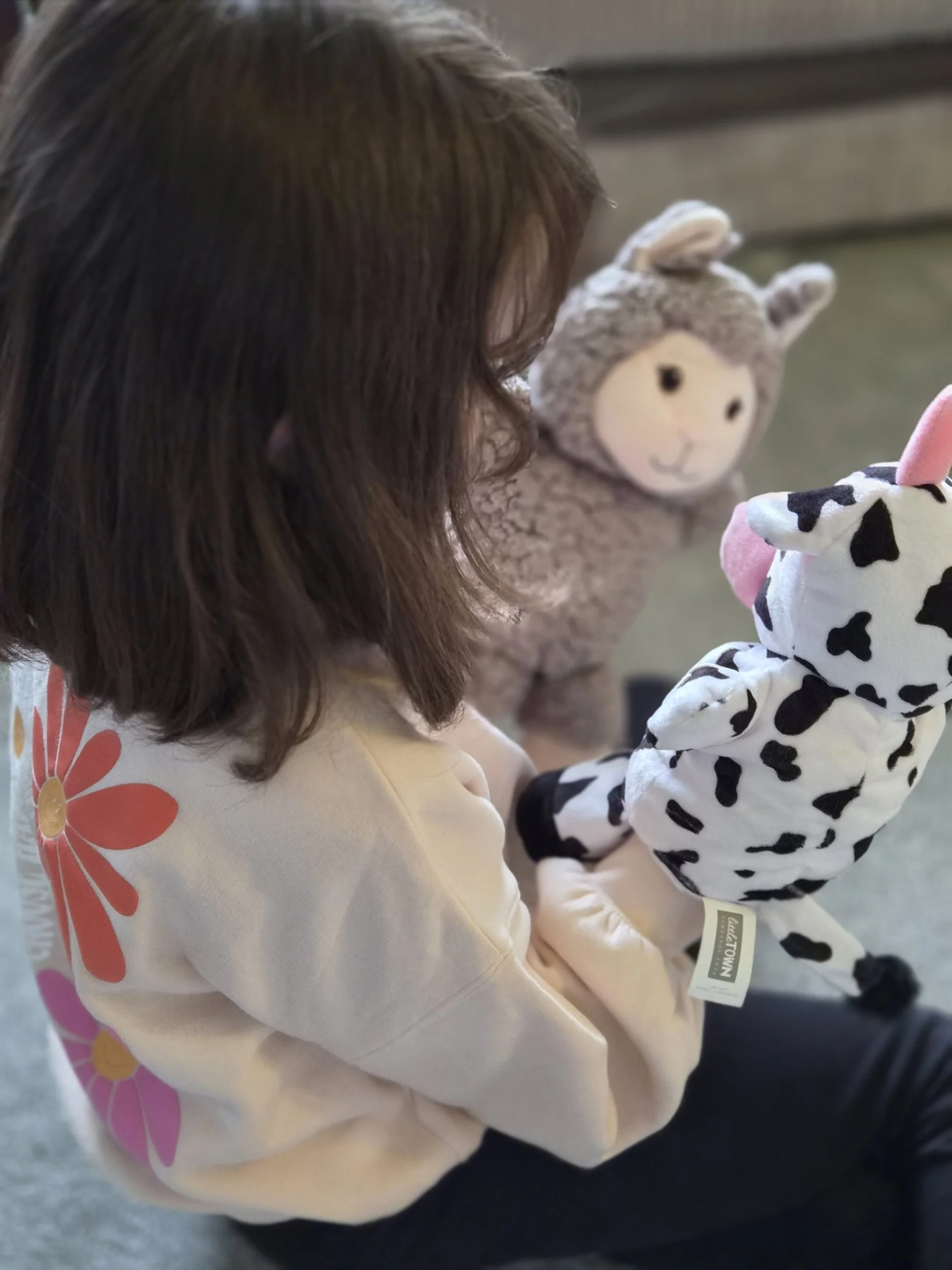 A young girl with brown hair is holding a stuffed animal, a cow-patterned plush toy, in her hands. She is wearing a cream shirt with pink and orange flower designs on the sleeve. The girl appears to be sitting, and the background shows a blurred indoor setting.