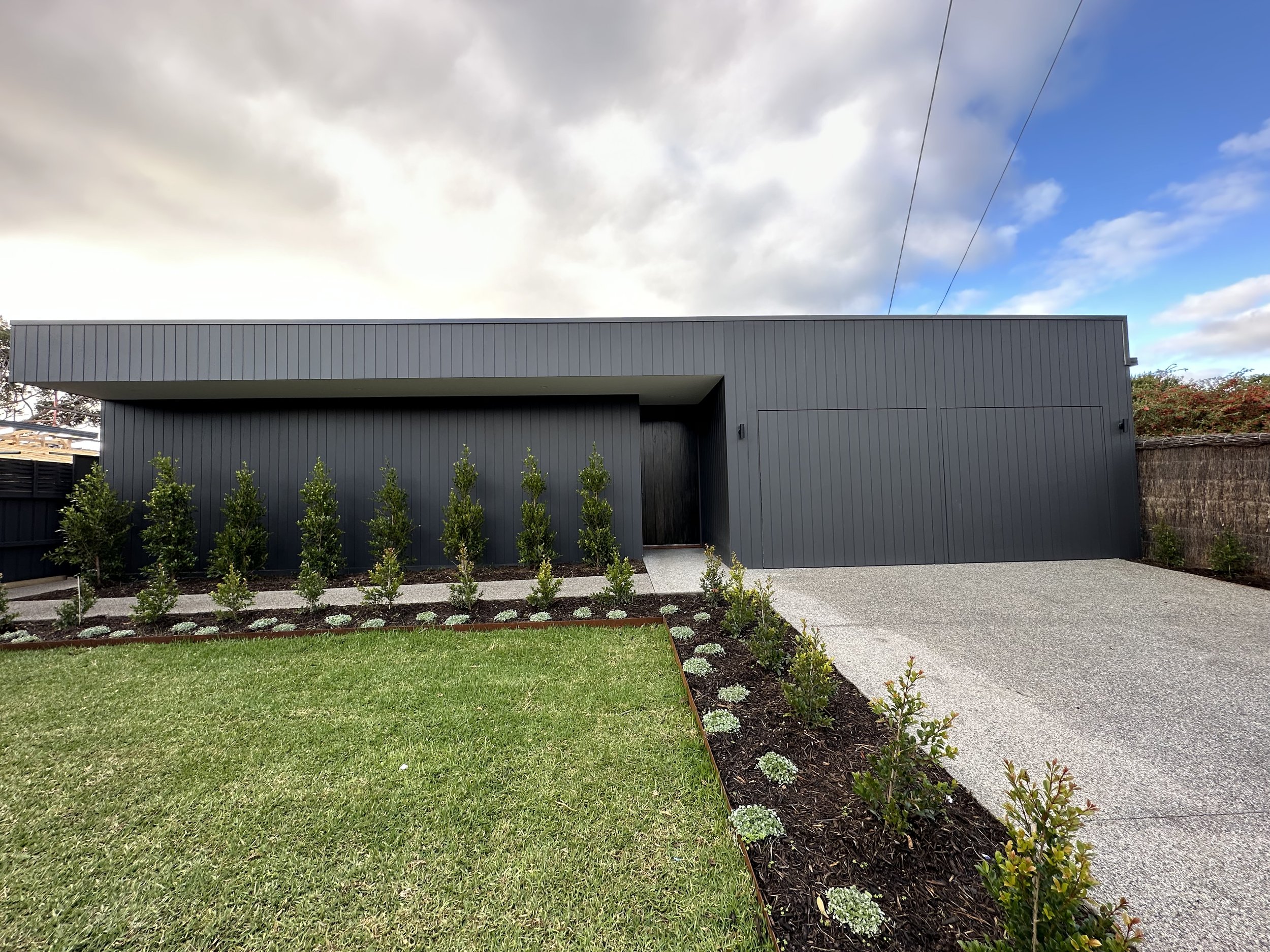 A modern, gray building with vertical metal siding, small windows, and a black door. In front, there is a well-maintained garden with green shrubs, a gravel pathway, a concrete sidewalk, and a green lawn. The sky above is partly cloudy with patches of blue.
