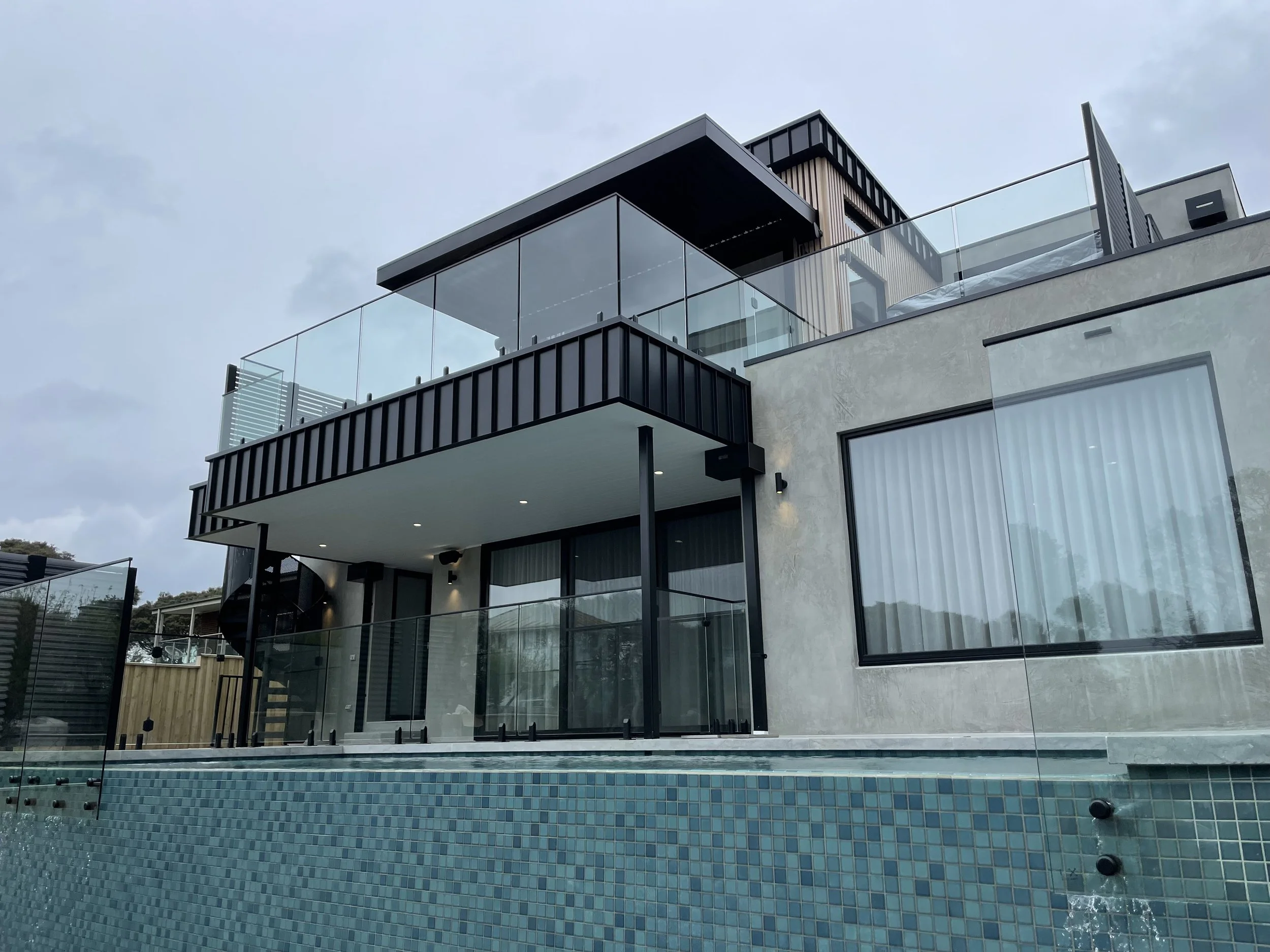 Modern multi-story house with large glass windows, black metal accents, and a rooftop terrace with glass railing, overlooking a swimming pool with a mosaic tile edge.