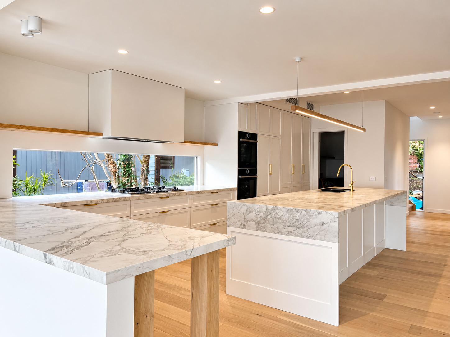 Modern kitchen with white cabinetry, marble countertops, and wooden accents. Features include a kitchen island with a gold faucet, built-in ovens, and large windows overlooking a garden.