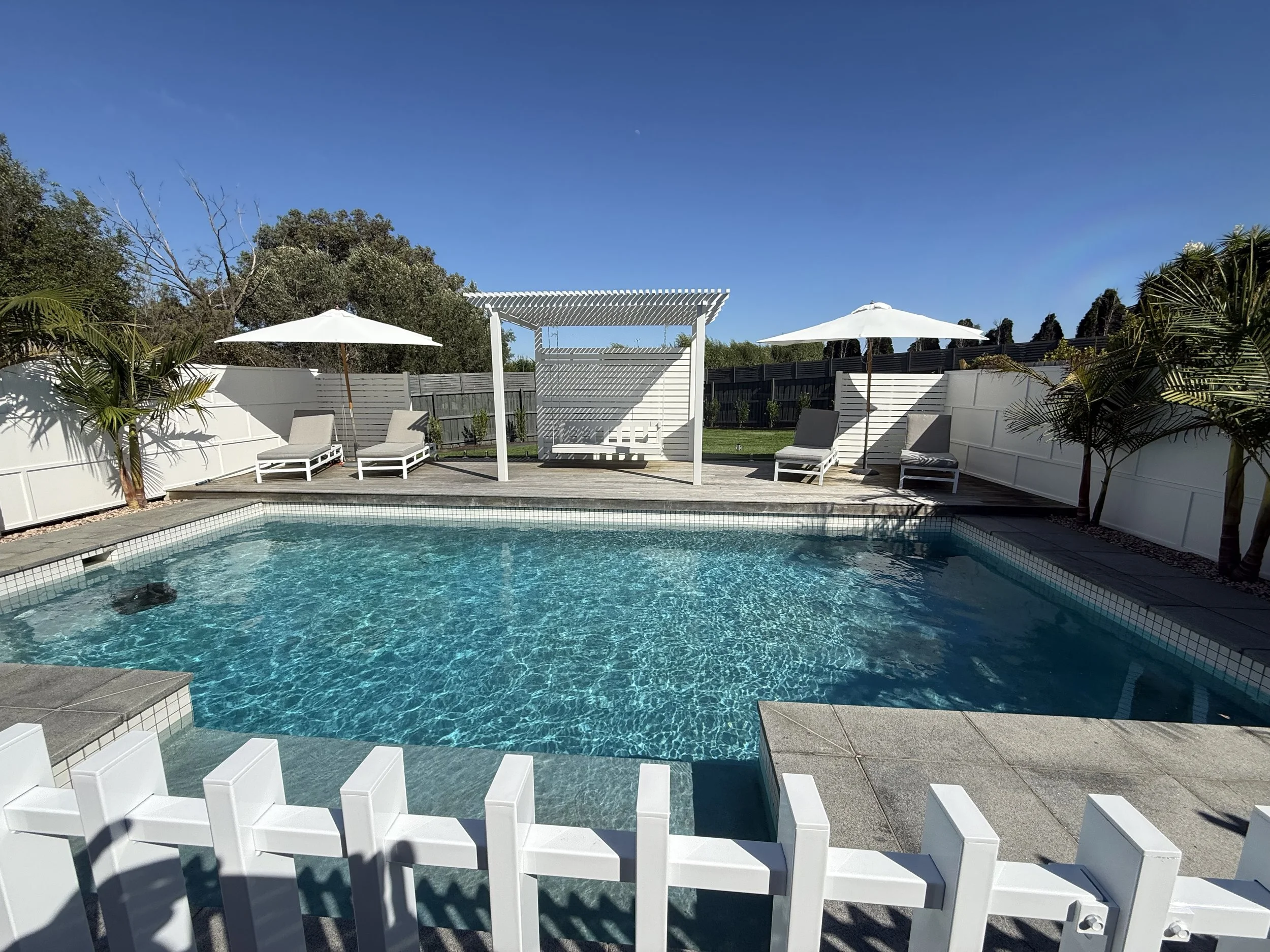 A backyard swimming pool with lounge chairs, white umbrellas, and a white privacy fence. There are some trees and bushes around the yard, and the sky is clear and blue.