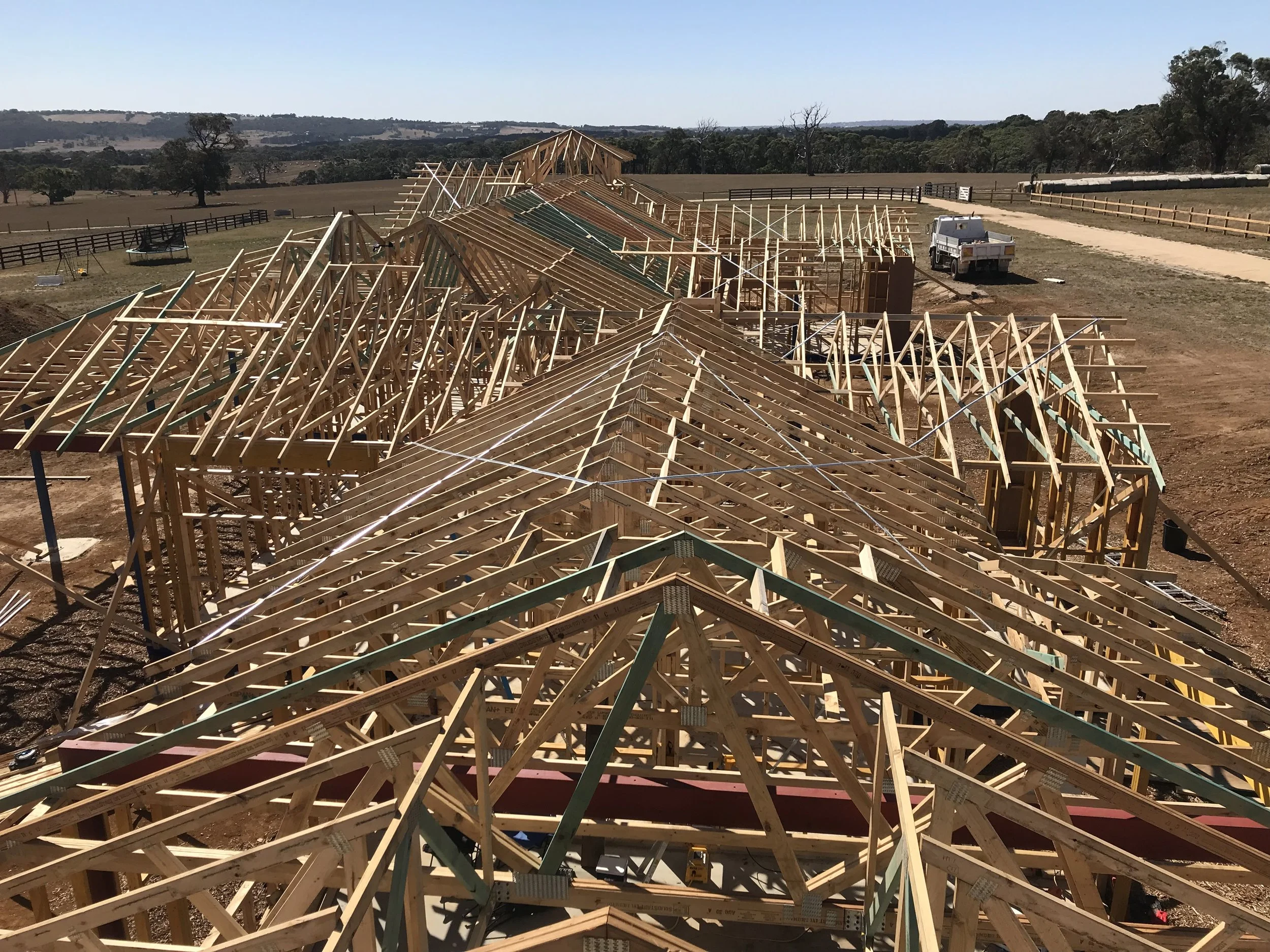 A large construction site of a house being built with a wooden roof framework, placed in an open field with trees and hills in the background.