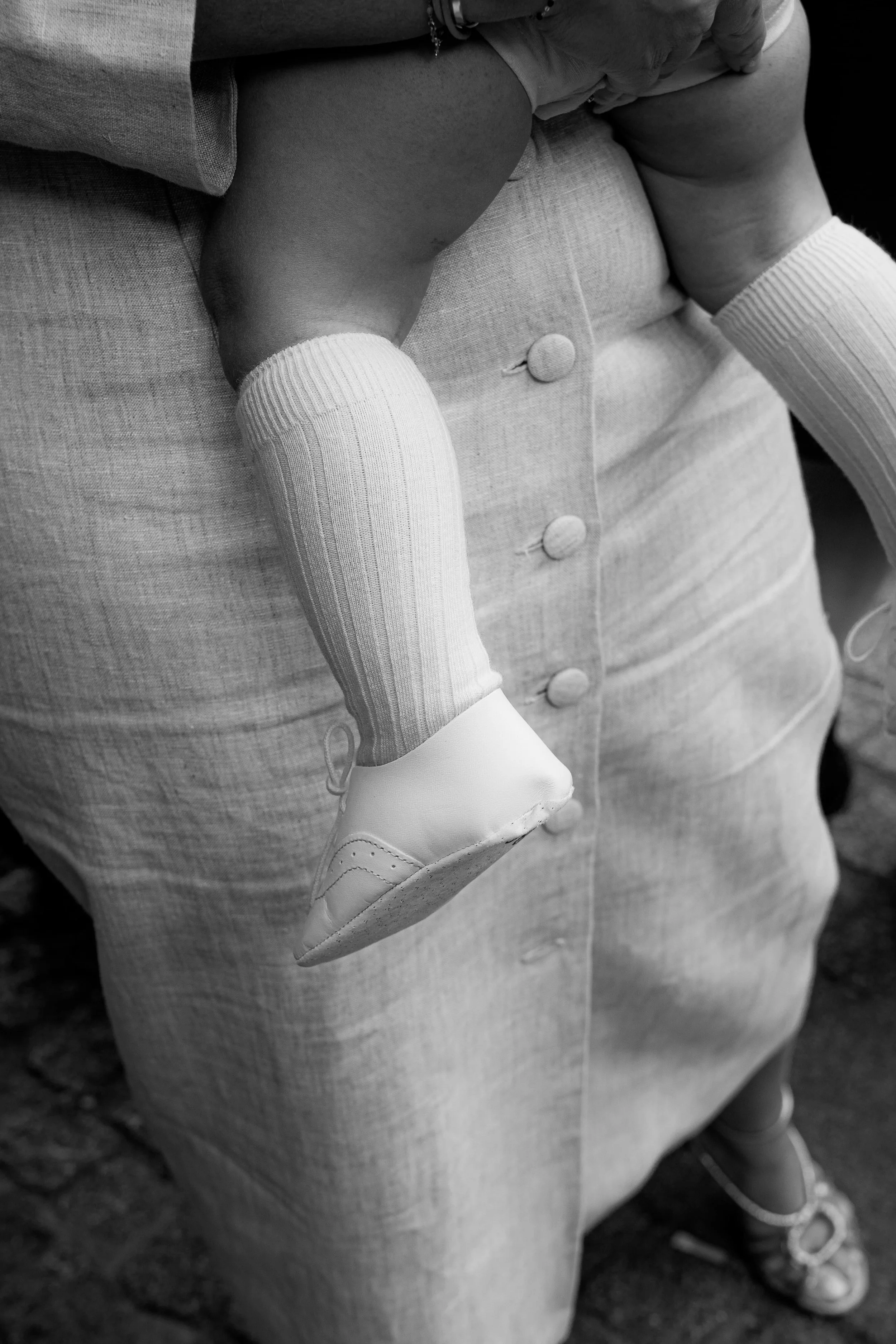 Un enfant assis sur les genoux d'une personne adulte, portant des chaussettes longues blanches, une robe ou un haut gris et une chaussure blanche. La photo est en noir et blanc.