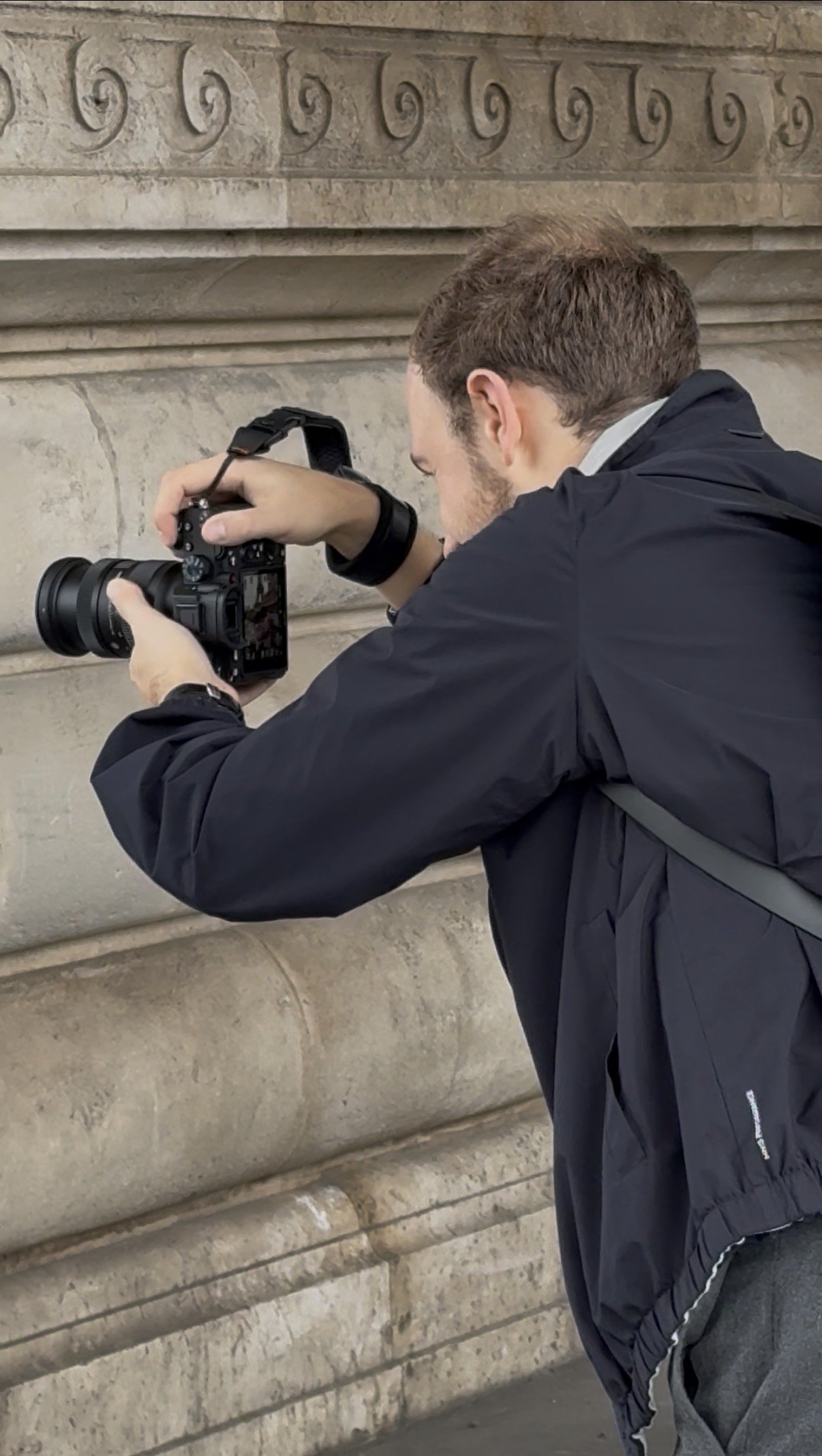 Un homme en veste noire prend une photo avec un appareil photo numérique contre un mur en pierre sculptée.