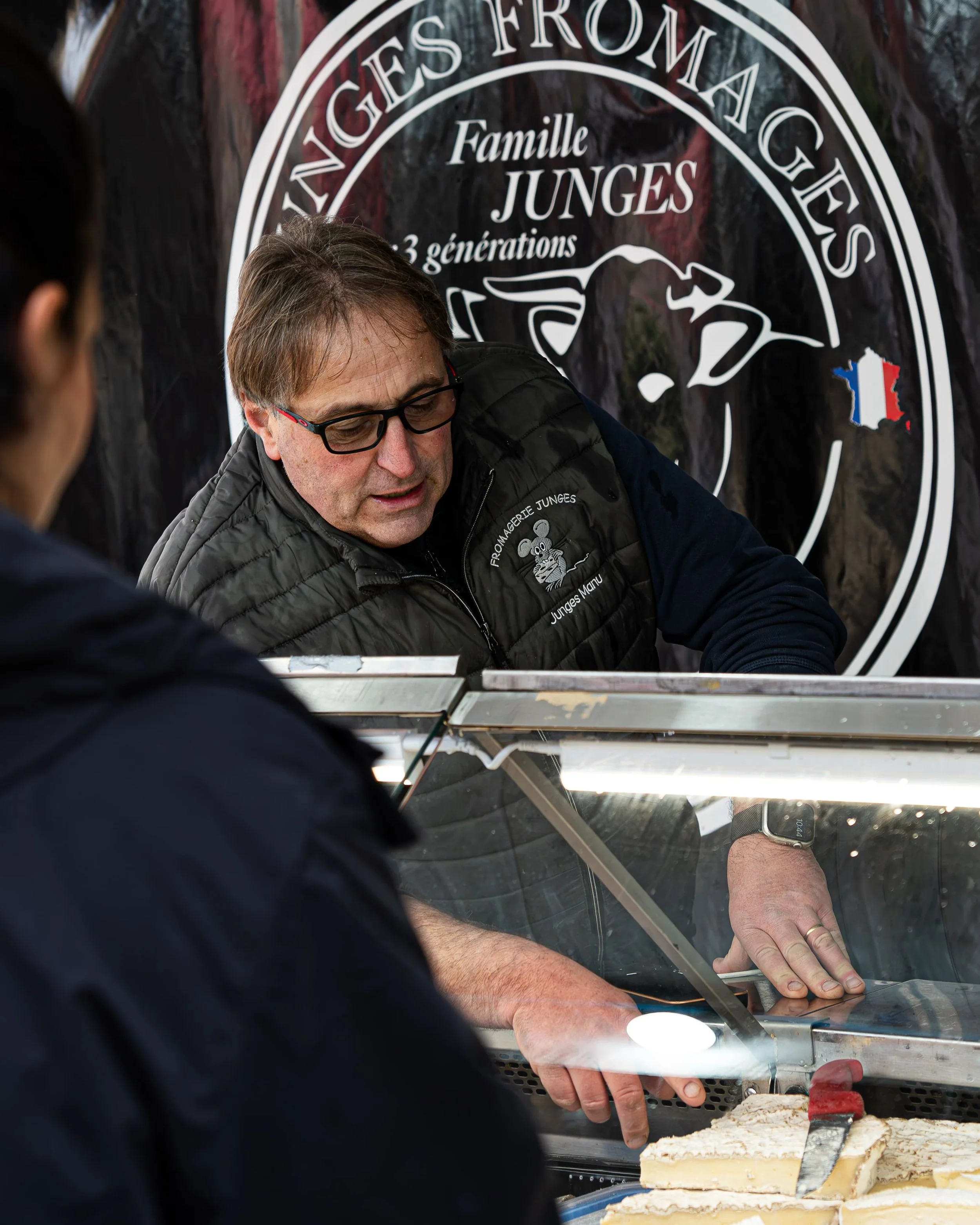 Deux hommes coupent une tarte ou un gâteau dans une vitrine, en arrière-plan un panneau avec le texte 'Vignes Fromages Famille Junges 3 générations'.