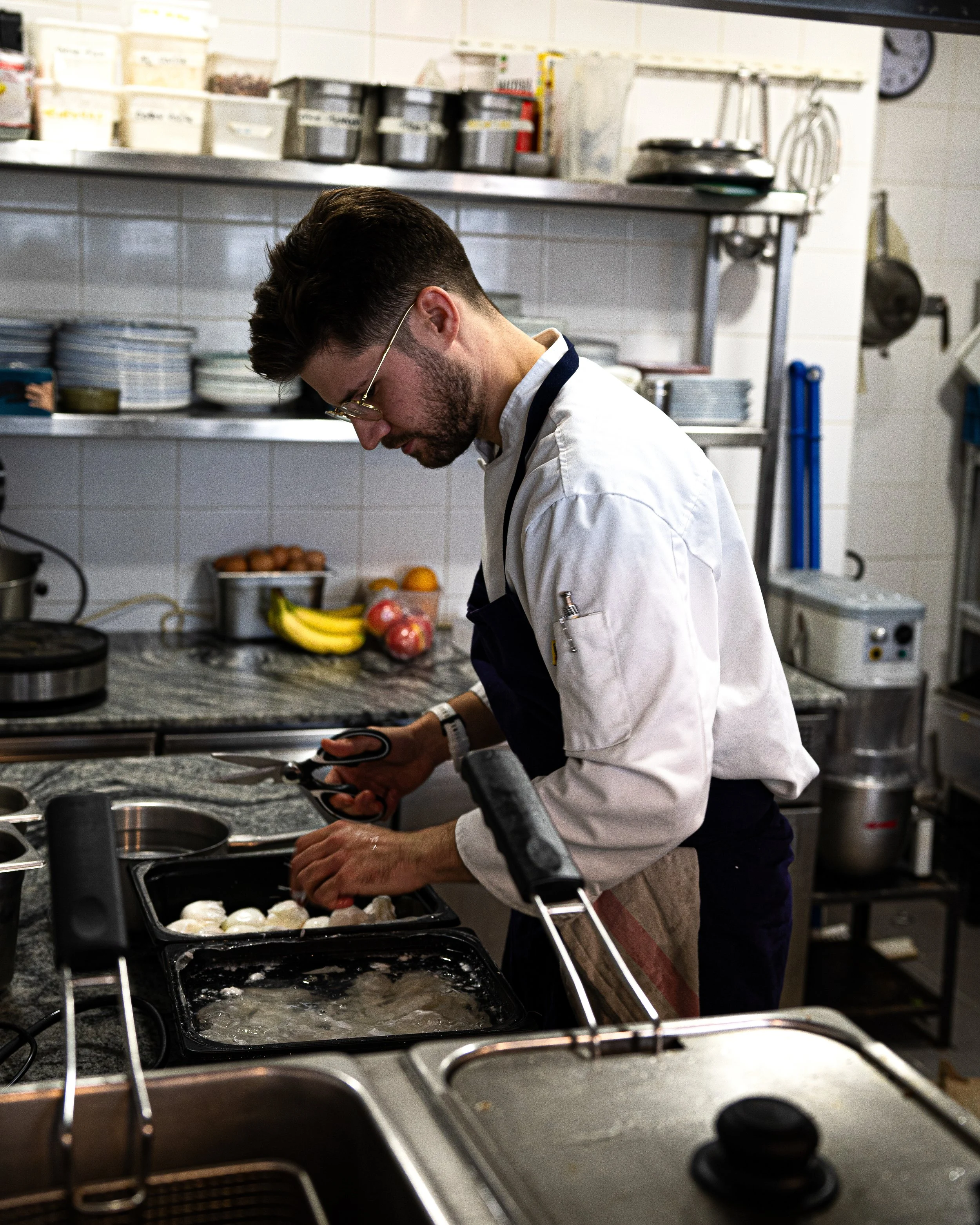 Un chef en cuisine préparant des aliments dans une cuisine professionnelle, avec des fruits, des légumes et des appareils de cuisine visibles.