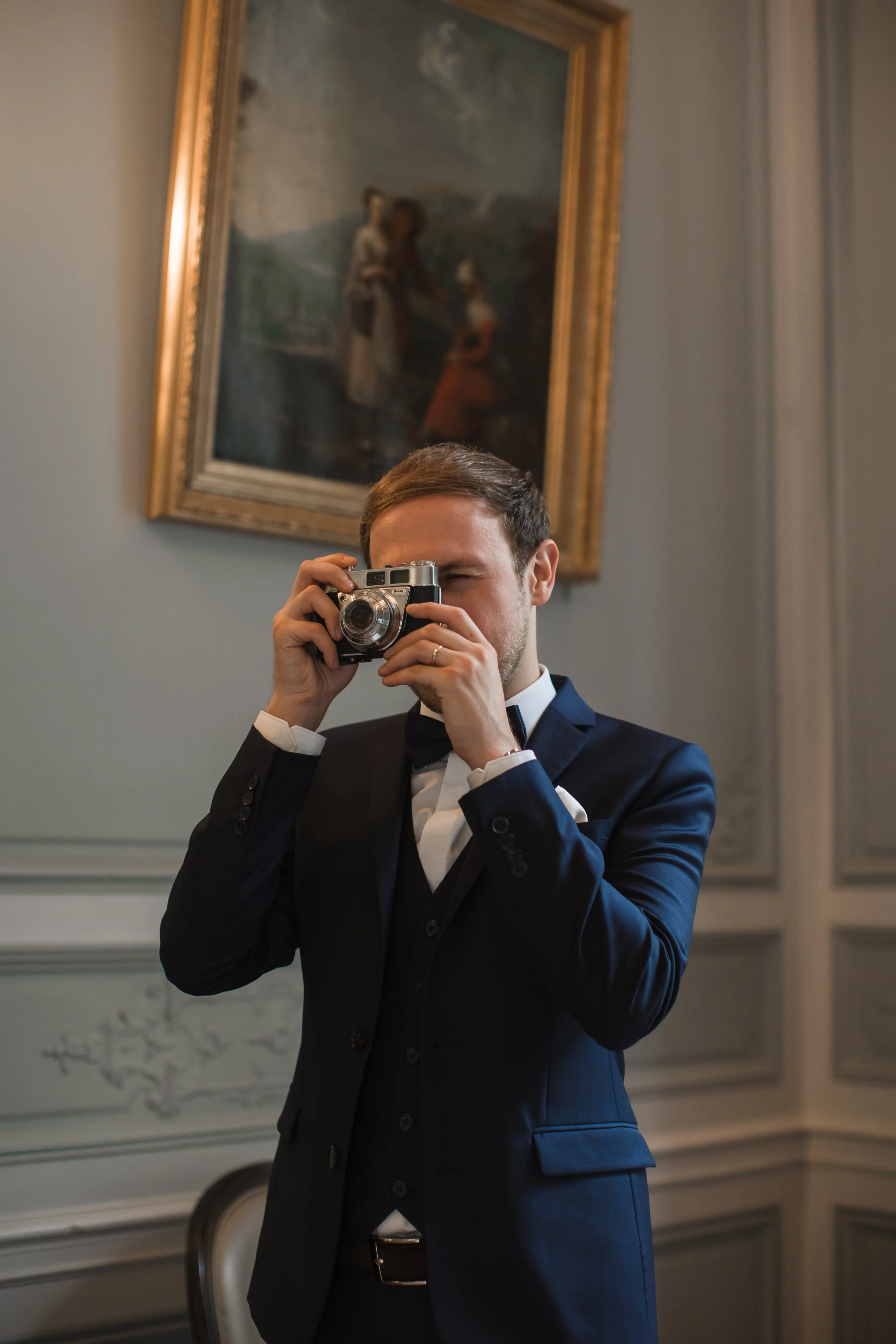 Un homme en costume noir, tenant un appareil photo devant son visage, dans une pièce élégante avec un tableau accroché au mur derrière lui.