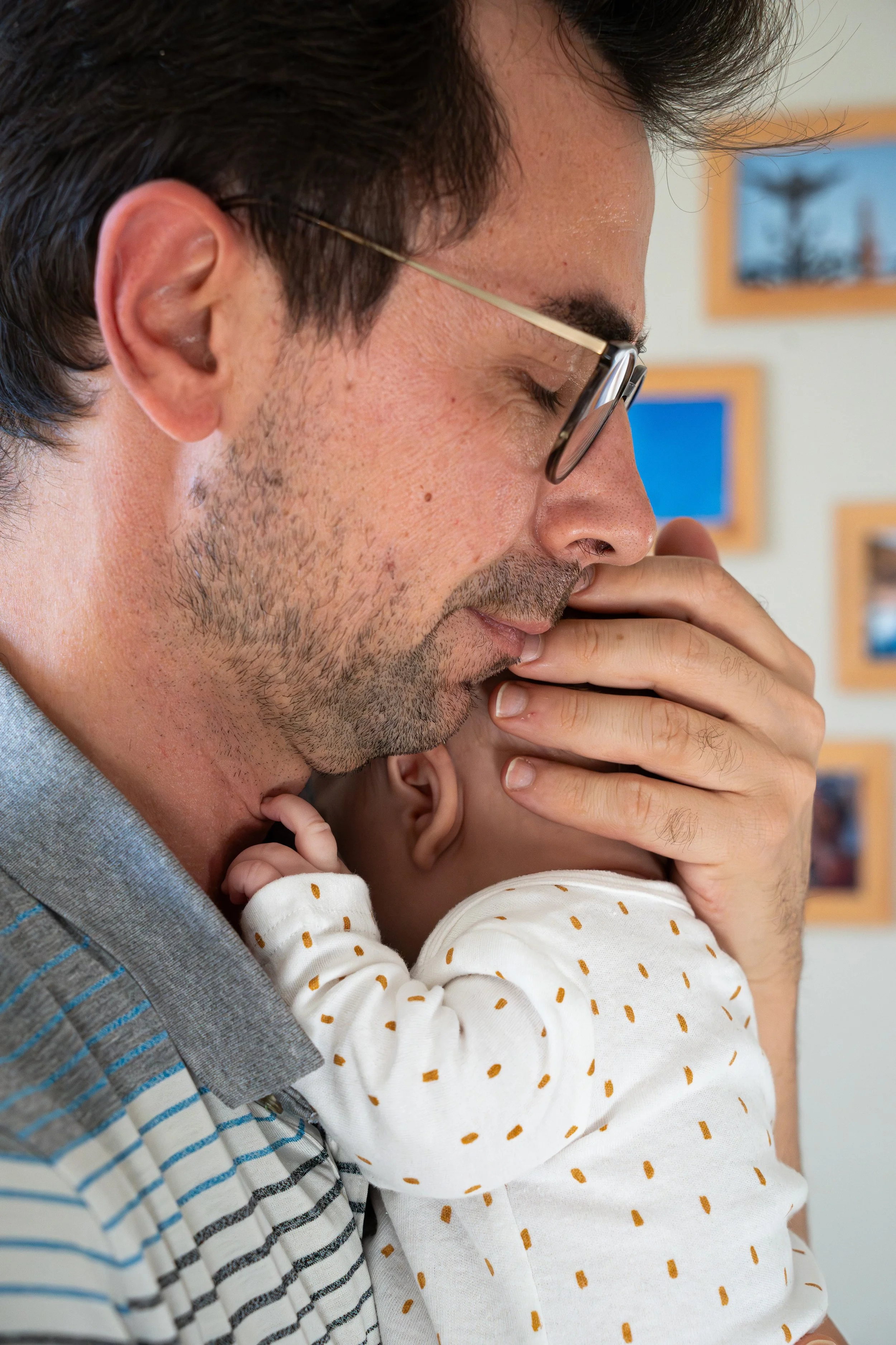 Un homme portant des lunettes tient un bébé dans ses bras, recomposé avec concentration. L'homme a des cheveux noirs et une barbe légère. Le bébé porte un pyjama blanc à points marron. Le fond comporte des cadres accrochés au mur.