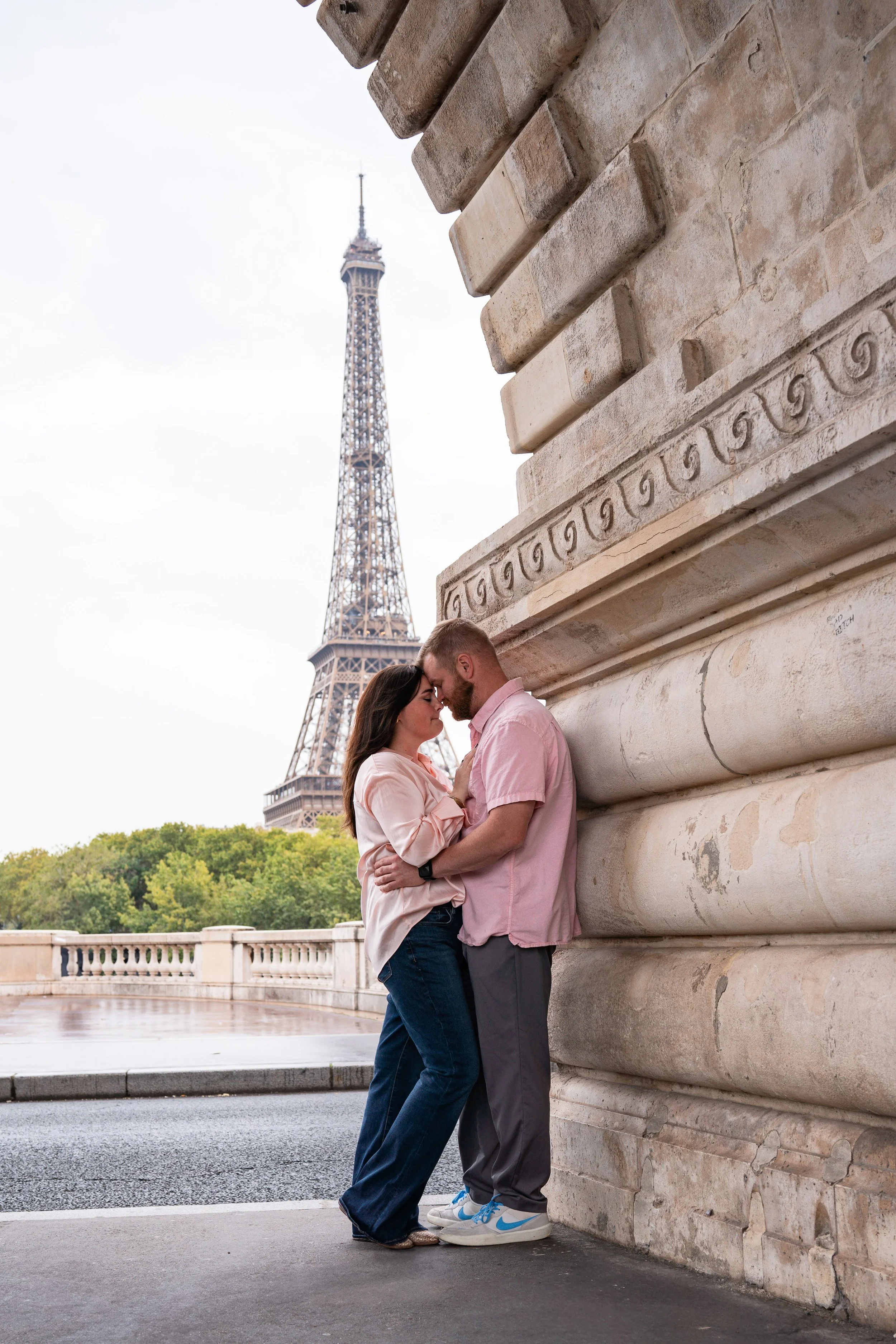 Un couple se tient très proche, les fronts se touchant, devant une structure en pierre à Paris avec la Tour Eiffel en arrière-plan. Le ciel est nuageux.