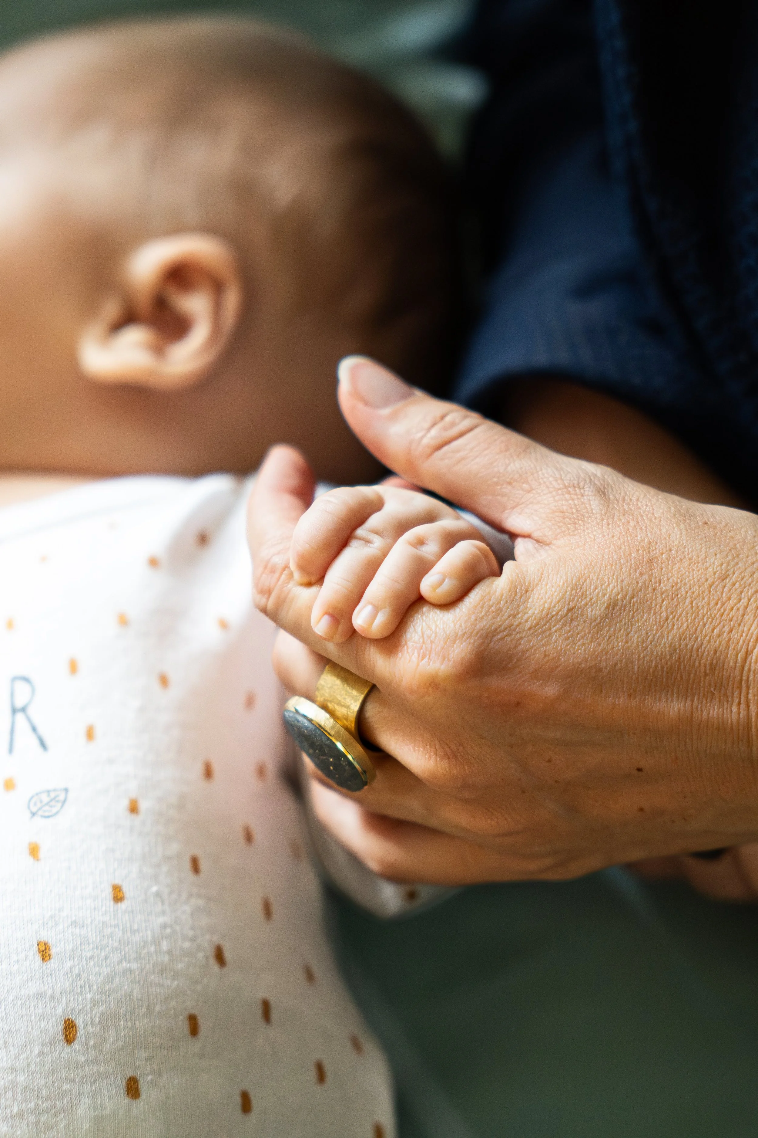 Une personne tient la main d'un enfant petit, probablement un bébé, dans une pièce. La main de l'adulte porte une bague noire avec une bande en or et une autre en argent. L'enfant porte un vêtement blanc avec de petits motifs tombant doucement.
